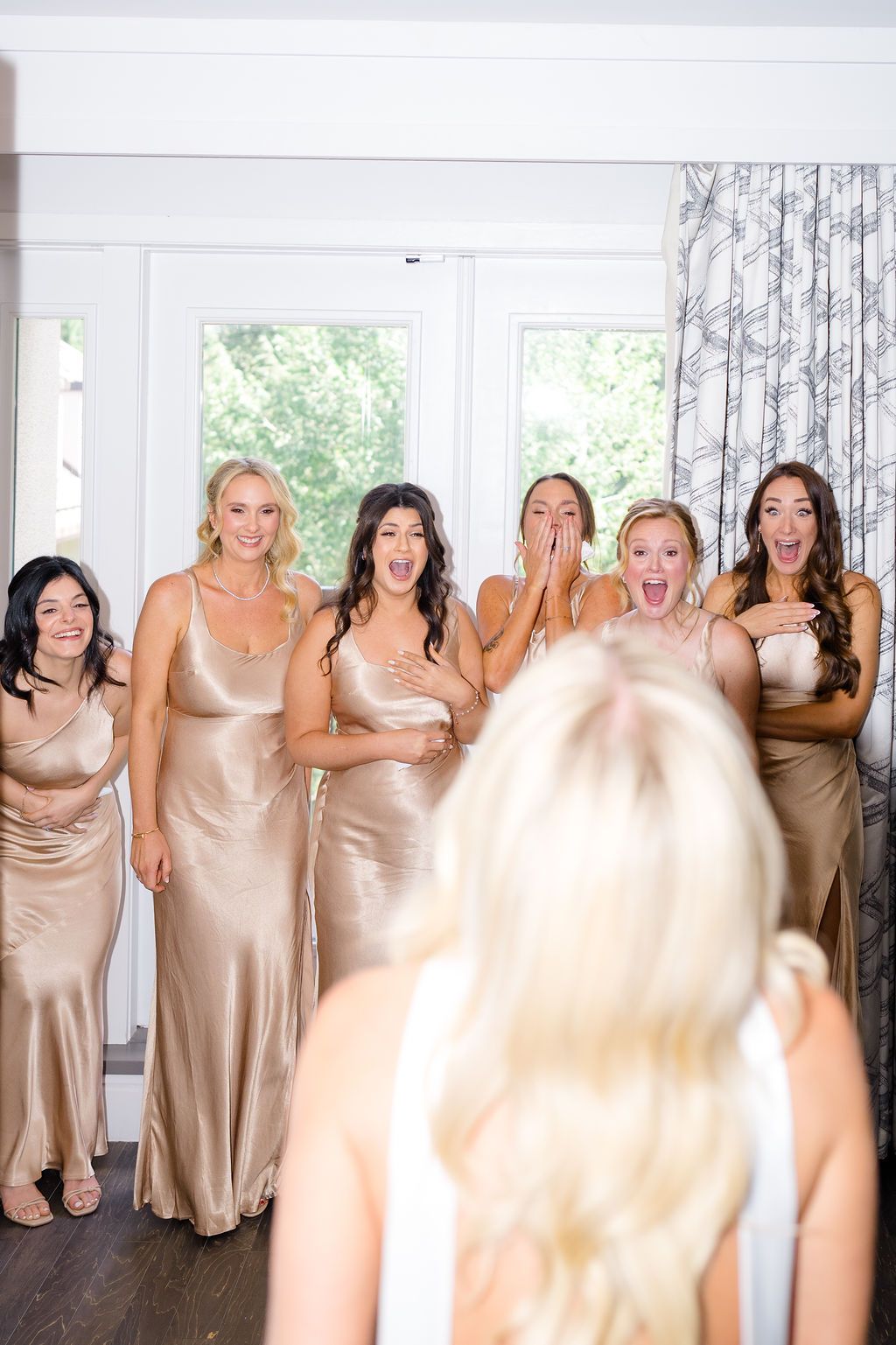 Bridesmaids in gold satin dresses react with joy and surprise seeing the bride in her wedding dress during bridal preparations.
