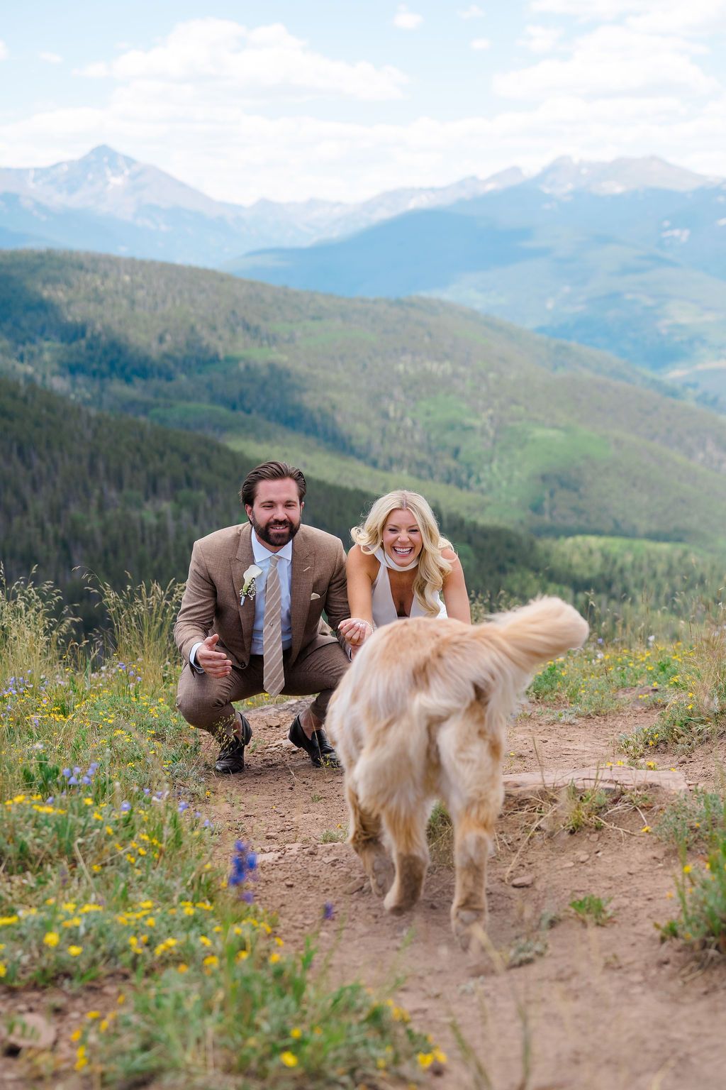Couple in wedding attire crouching with golden retriever on mountain trail with scenic Colorado peaks backdrop.