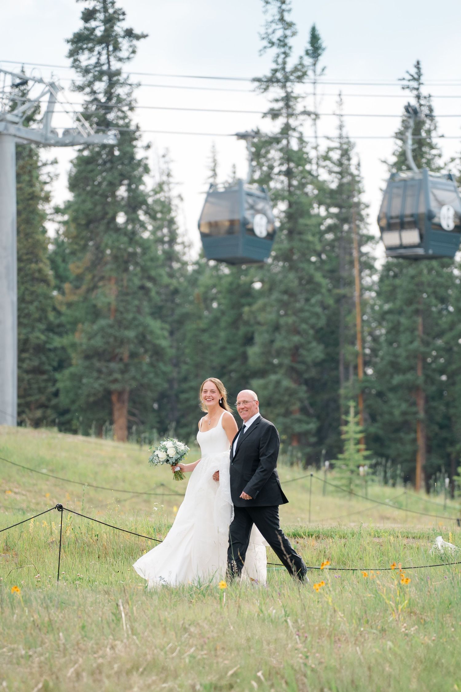 Wedding couple walks down grassy hillside at mountain resort with evergreen trees in background at copper mountain aerie venue.