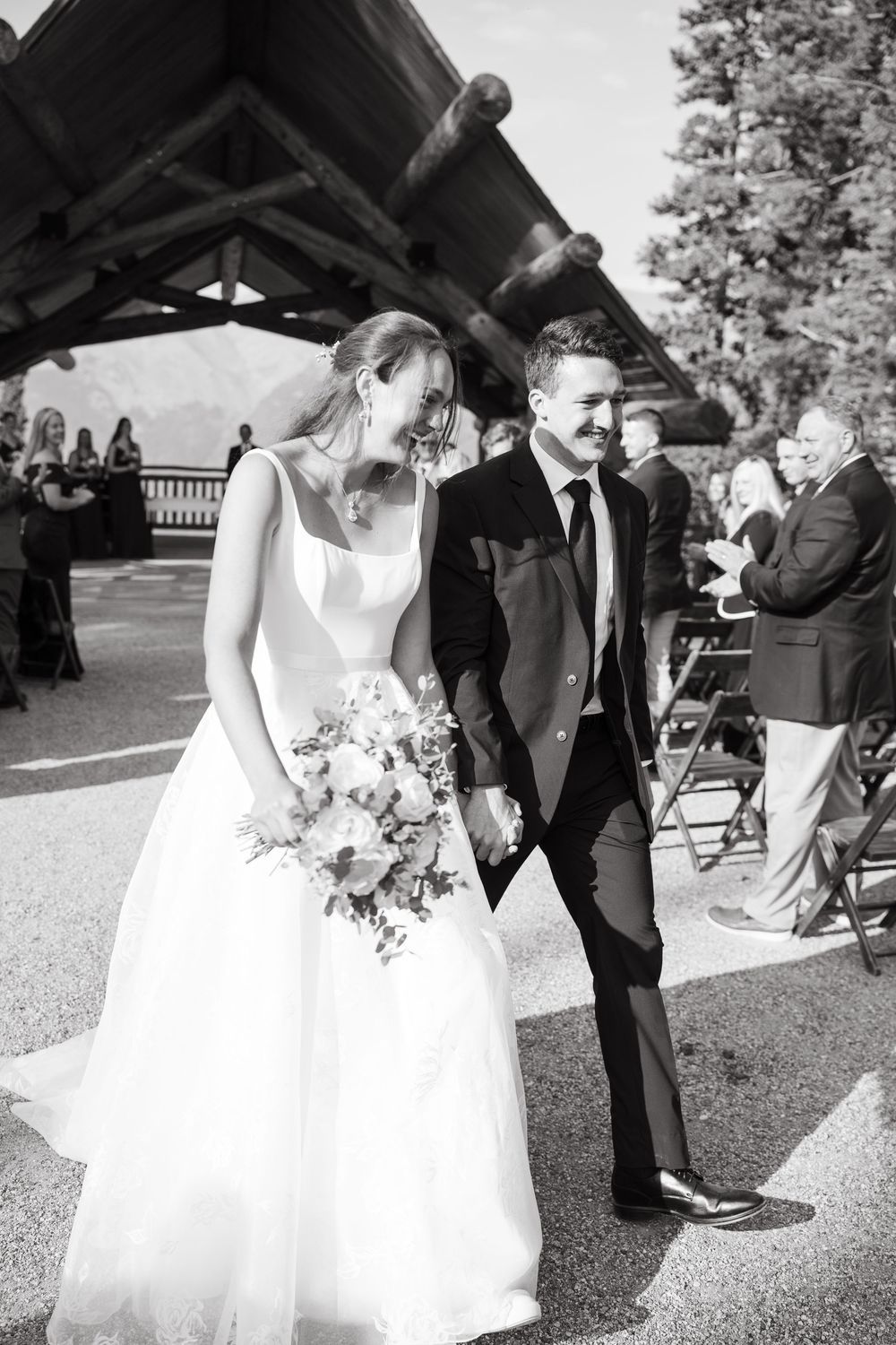 Black and white photo of a smiling bride and groom walking hand in hand down an outdoor wedding aisle past seated guests.