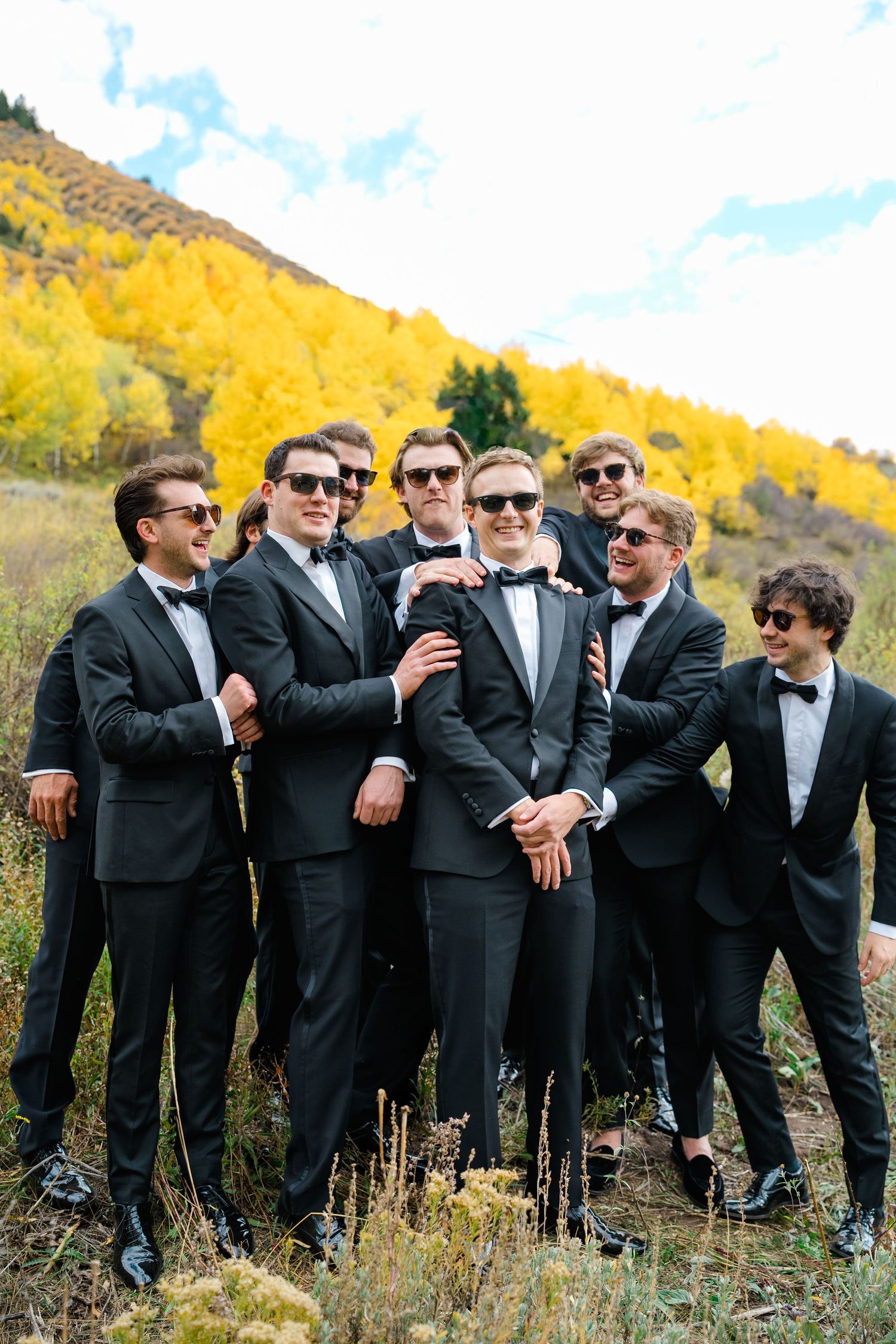 A group of groomsmen in black tuxedos and sunglasses pose outdoors with golden autumn aspen trees in the background.