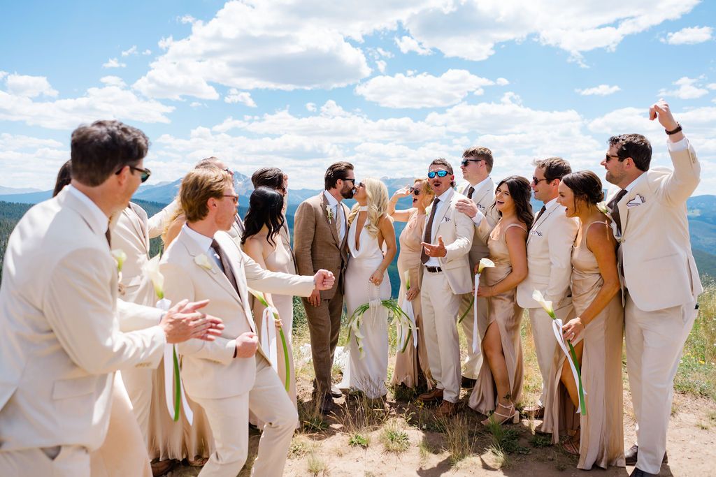 Wedding party in neutral attire celebrating outdoors on a sunny hilltop with scenic mountain views.