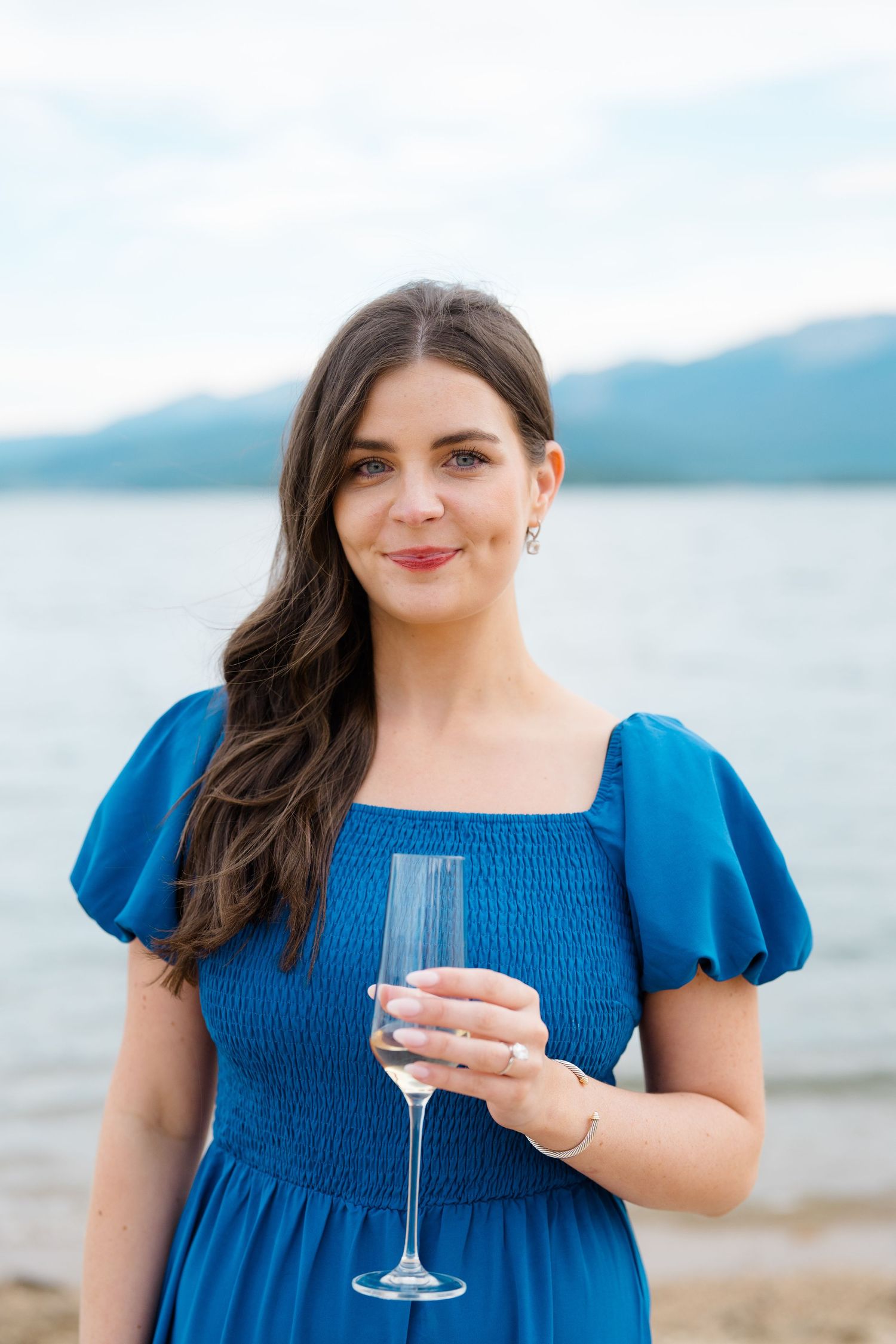 A woman in a blue dress stands by a lake holding a wine glass against a mountain backdrop at lake dillon.