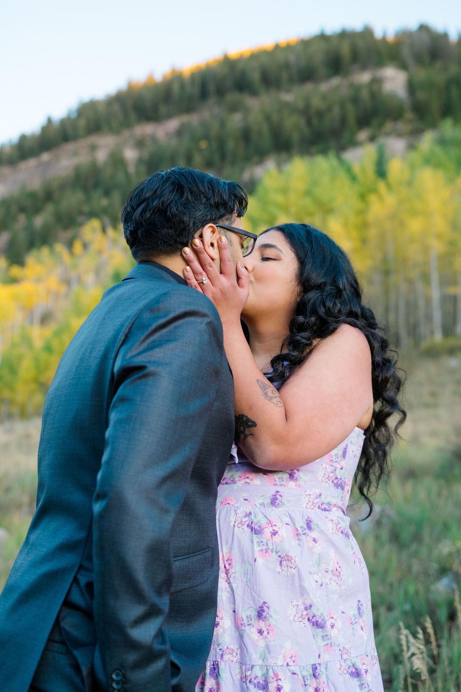 A romantic couple shares a kiss outdoors with autumn foliage and mountains in the background.