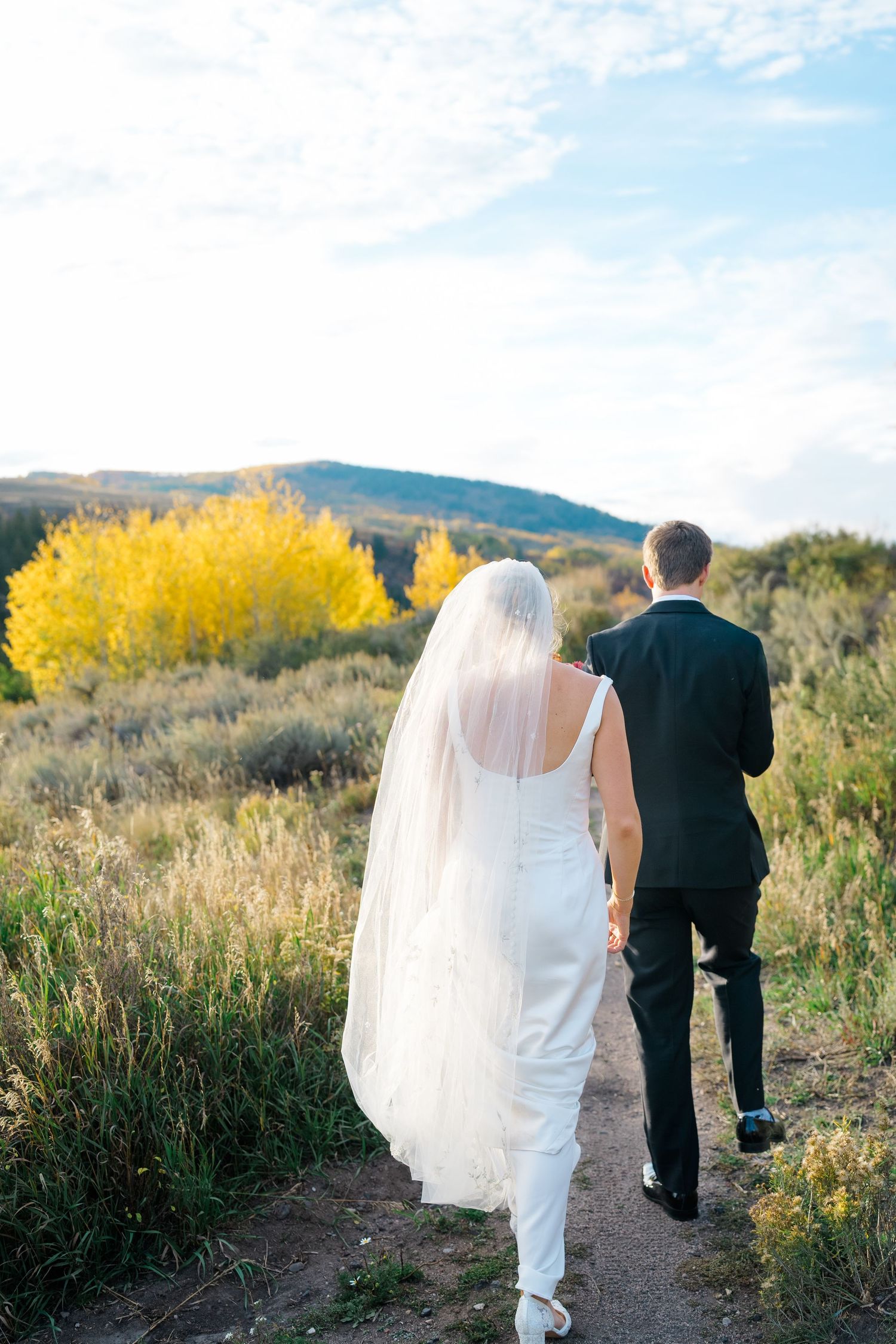 A couple in wedding attire walks along a scenic mountain trail with autumn foliage in the background.