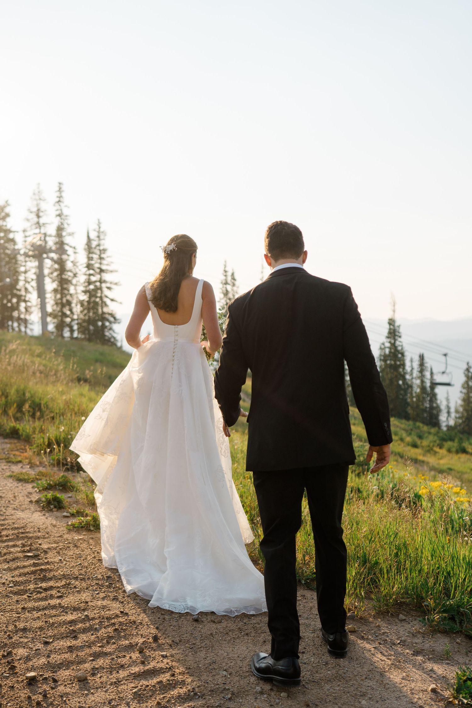 Wedding couple walking hand in hand along a mountain trail surrounded by evergreen trees.