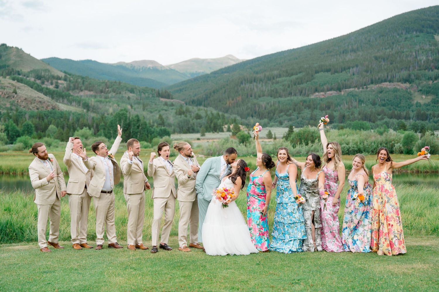 Wedding party in colorful floral dresses and khaki suits poses against scenic mountain backdrop in Colorado.