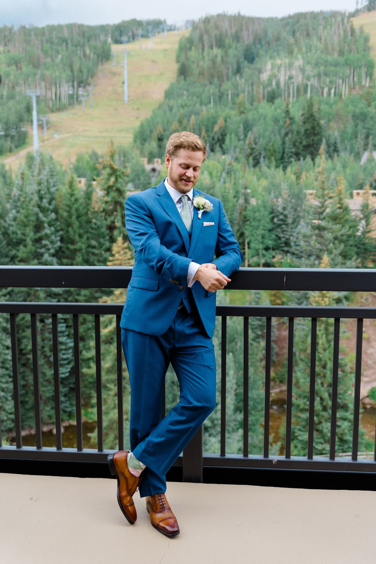 Groom in bright blue suit sits casually on outdoor deck railing with mountain forest landscape in background.