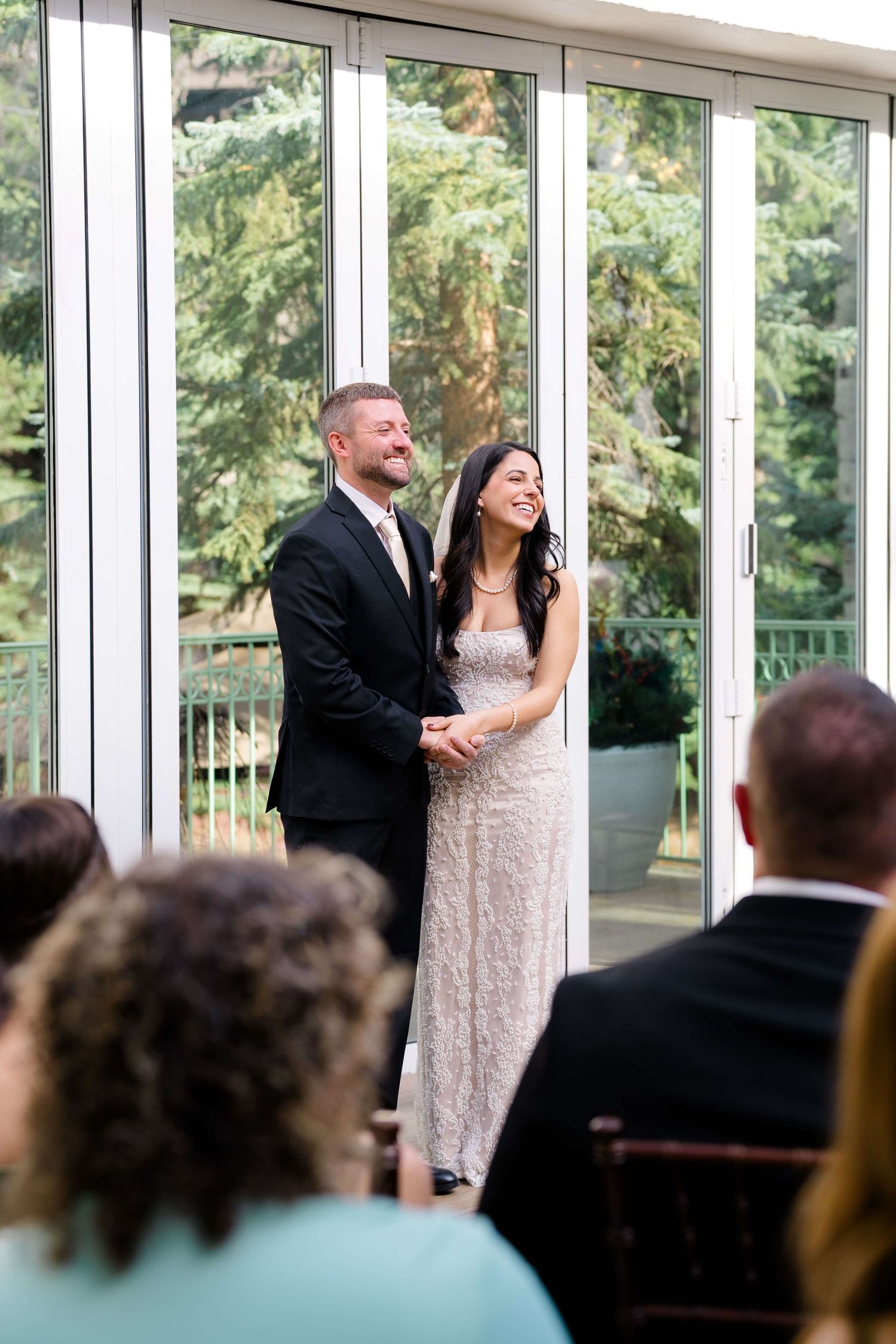 Bride and groom laughing joyfully at wedding altar in elegant glass venue with lush greenery backdrop.