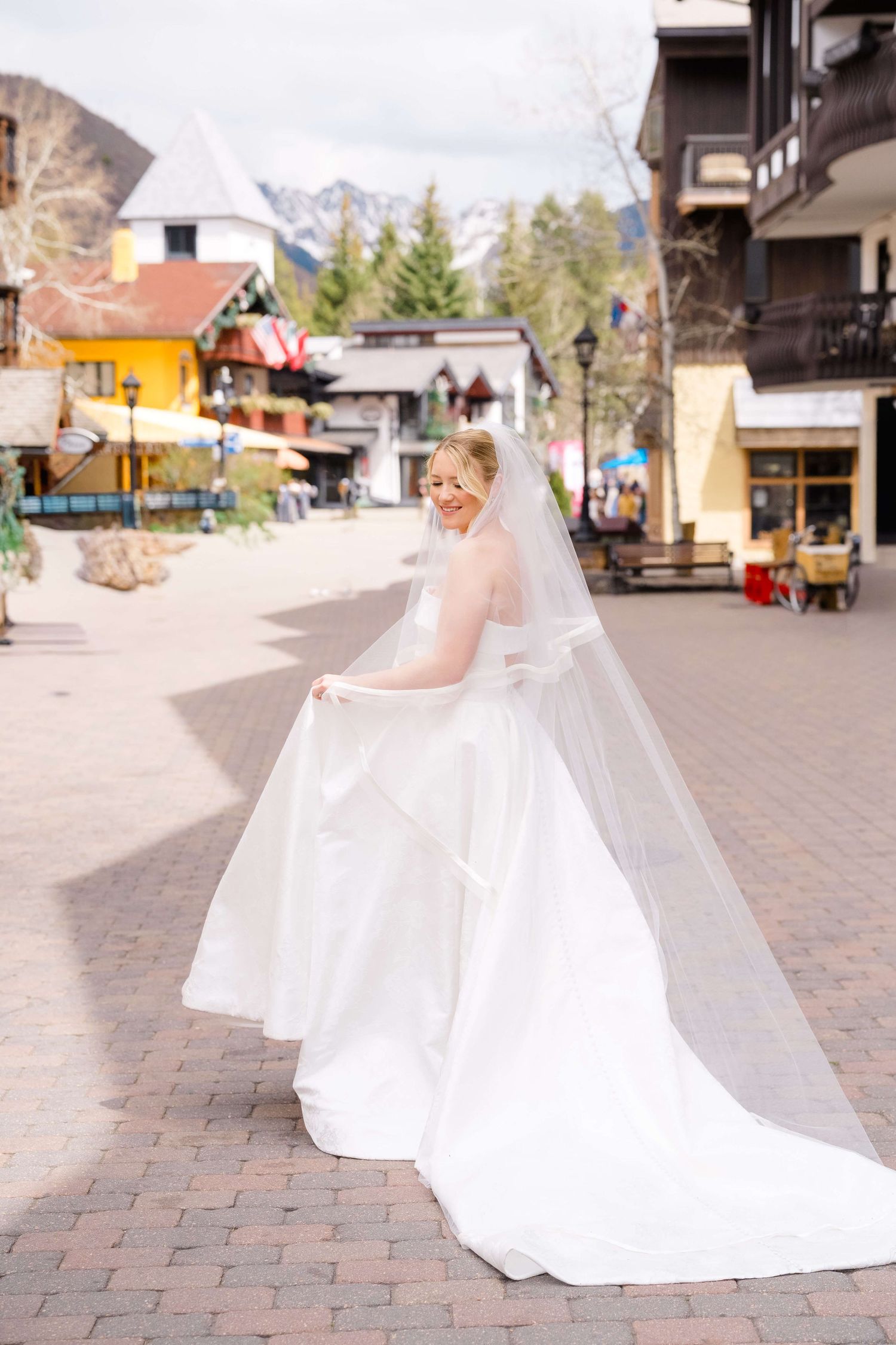 Bride in white ballgown and veil twirling in a charming mountain village square with snow-capped peaks.