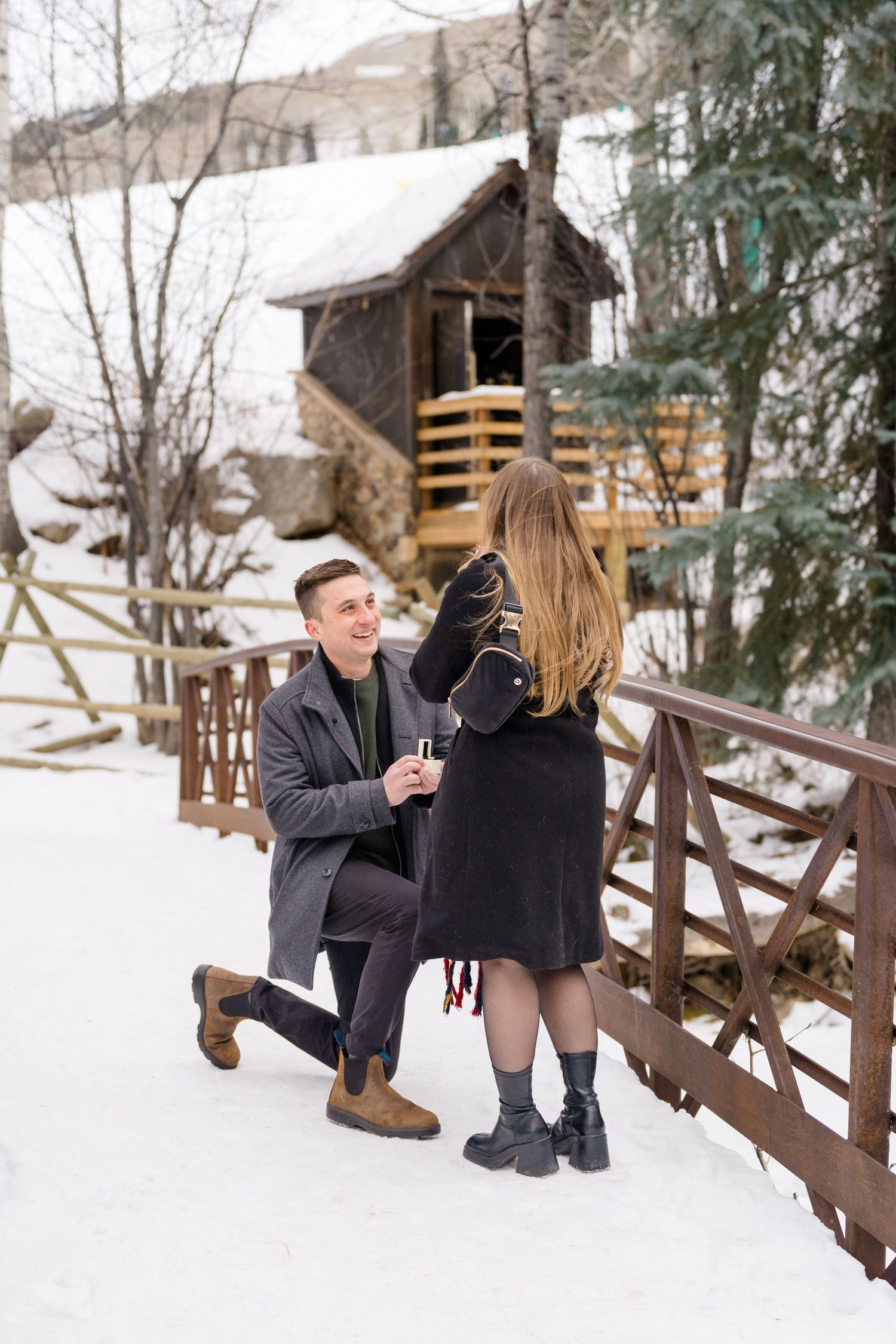 A couple sits together on a rustic wooden fence in a snowy winter landscape with evergreen trees in the background.