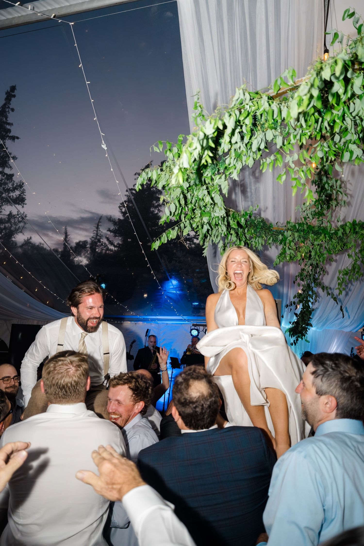 Bride laughing joyfully while being lifted on chair during hora dance at elegant tented wedding reception.
