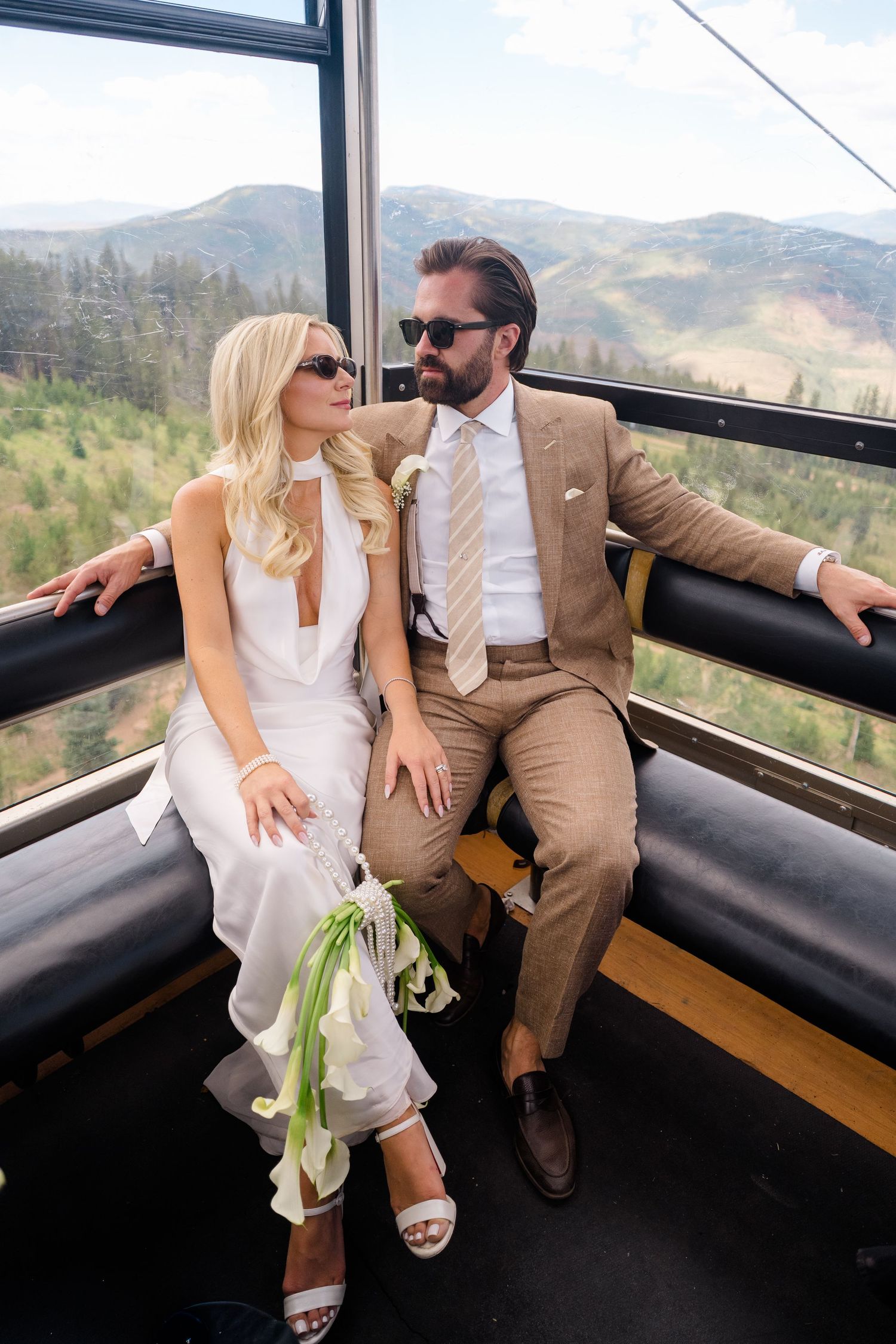 Bride and groom in wedding attire ride a gondola with mountain views, she in white dress, he in tan suit.