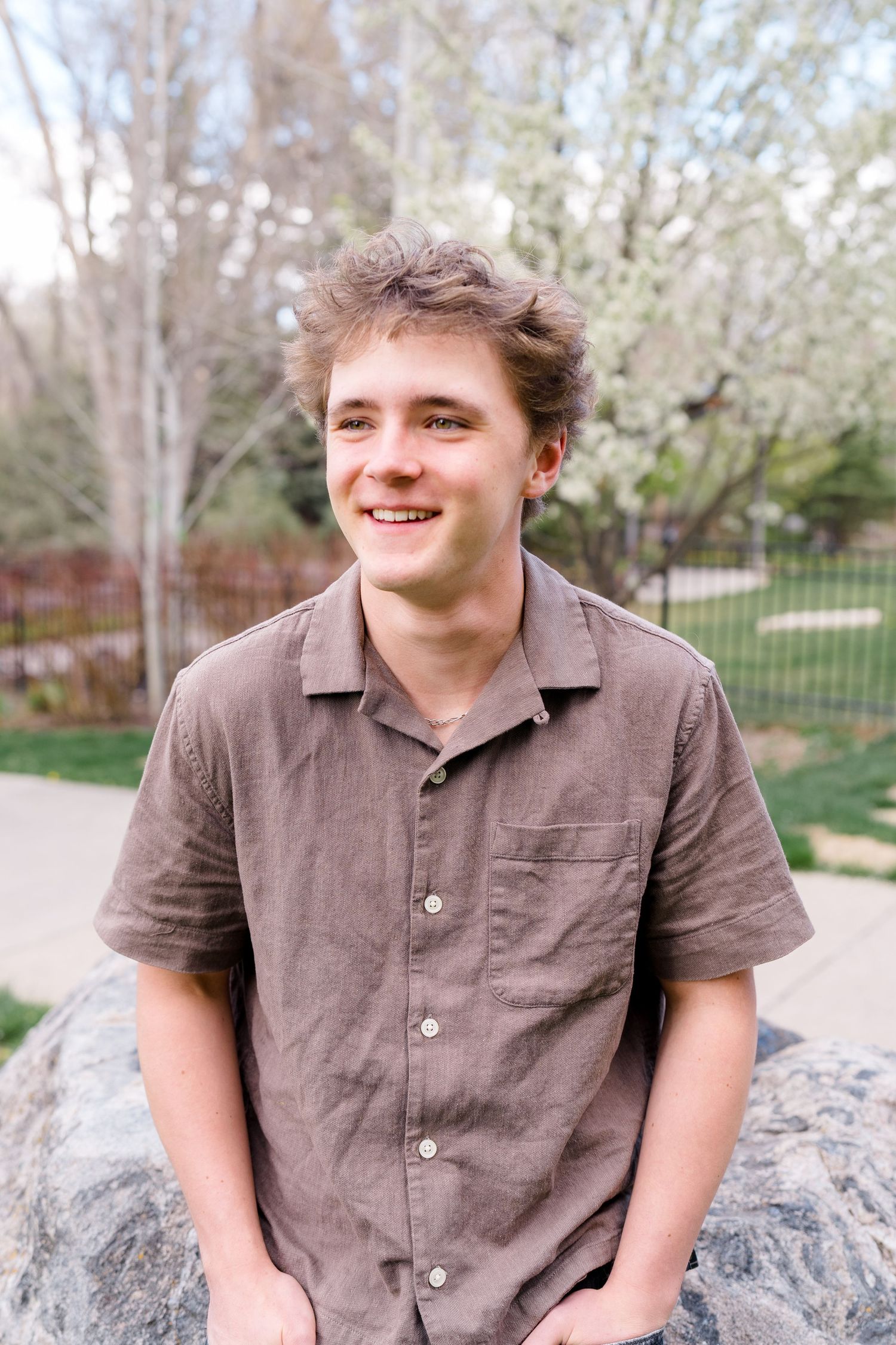 Smiling young man in a brown linen shirt sitting on rocks outdoors with blooming trees in the background.