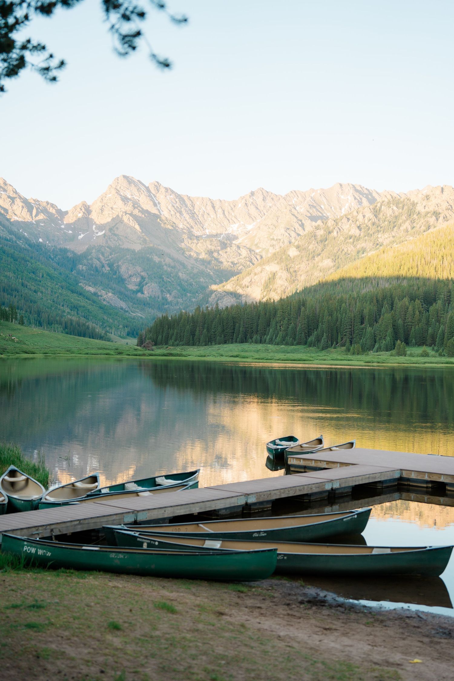 Green canoes docked at a wooden pier on a calm mountain lake surrounded by pine forests and rocky peaks at sunset.