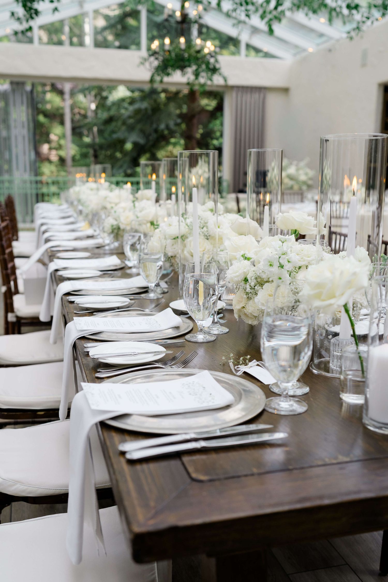 Elegant wedding reception table with white floral centerpieces, candles, and white linens on a rustic wood table.