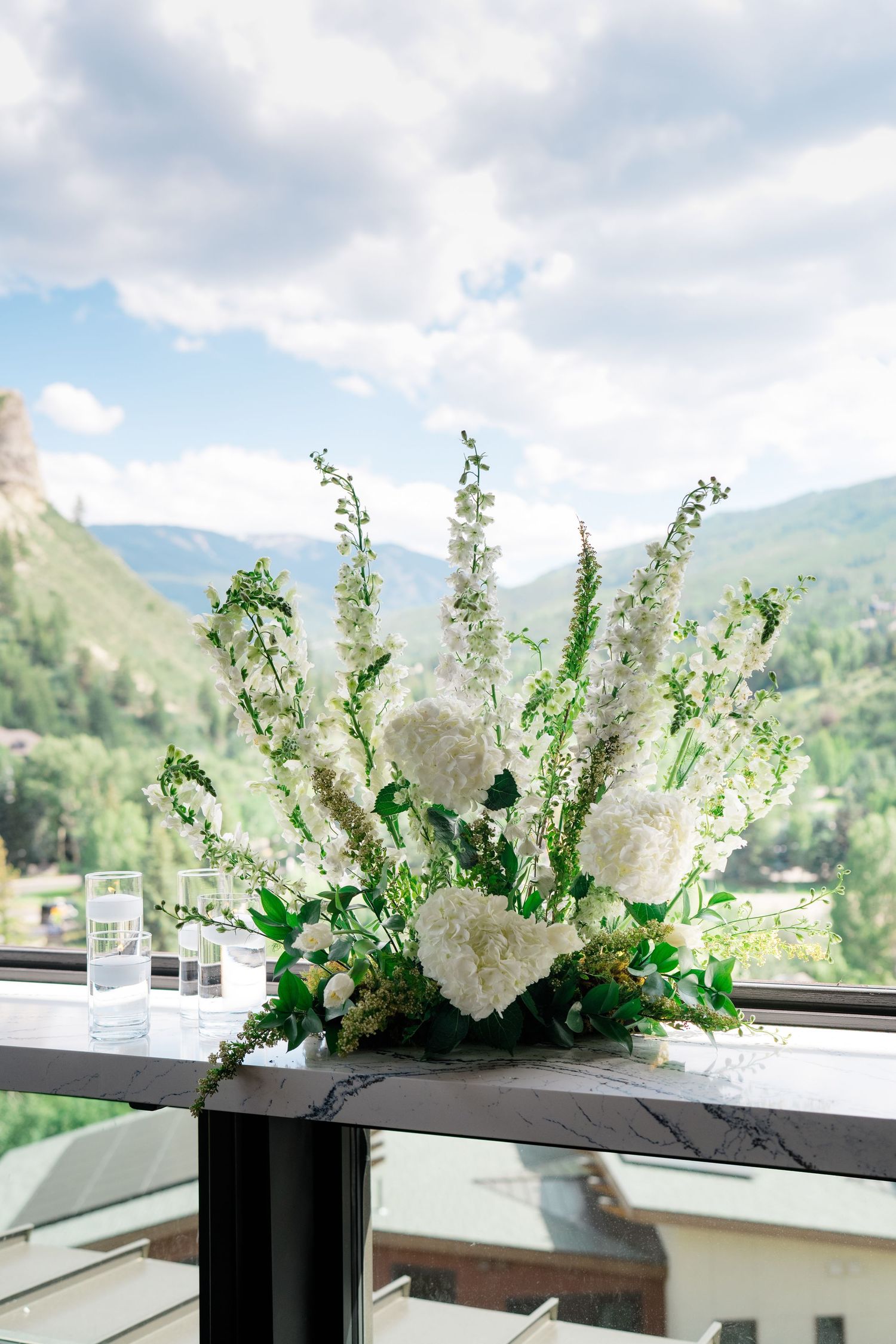White floral arrangement with hydrangeas and delphiniums on a balcony railing with mountain views.