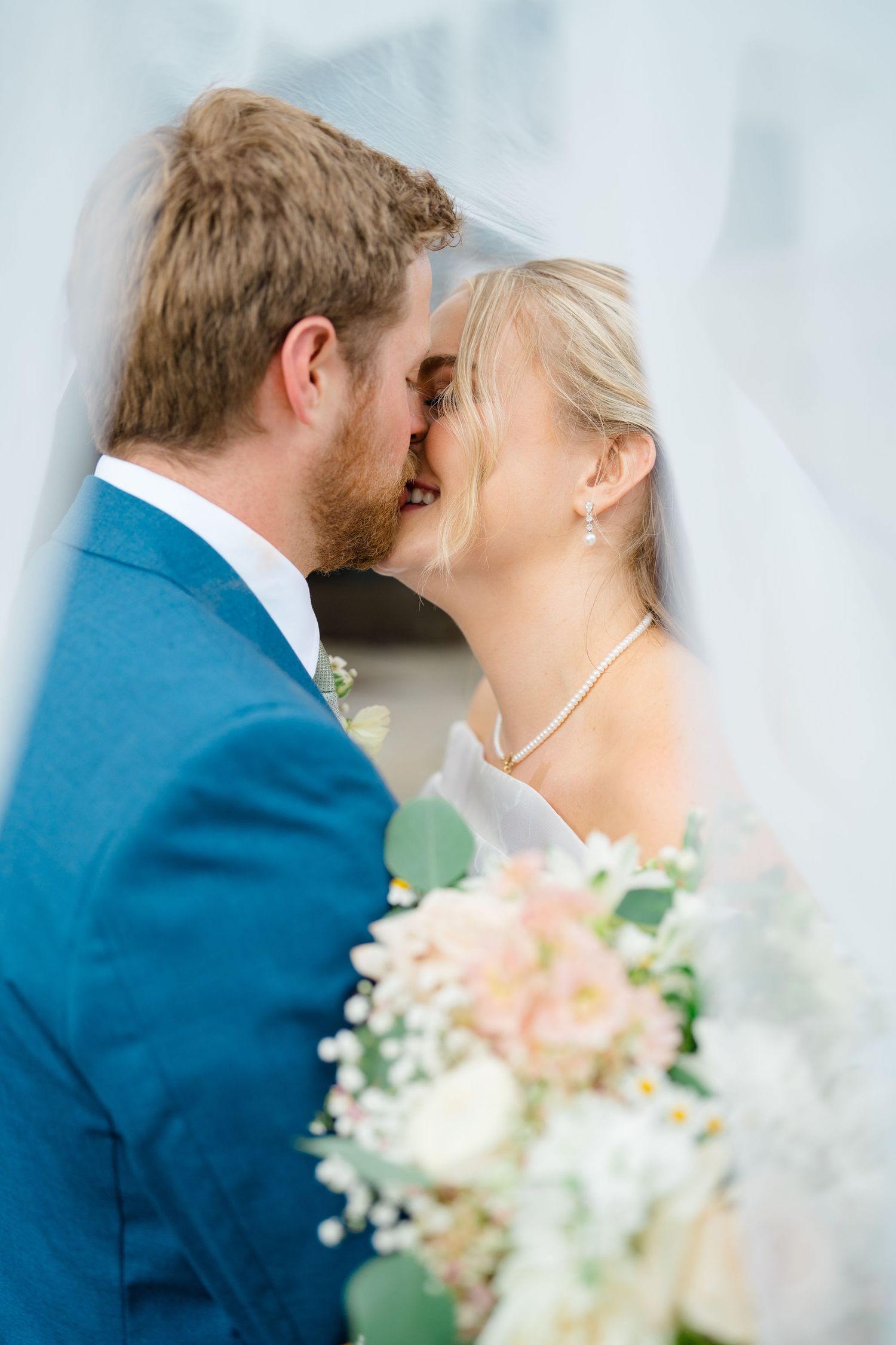 A couple in wedding attire share a romantic moment with soft pink and white flowers in the foreground.