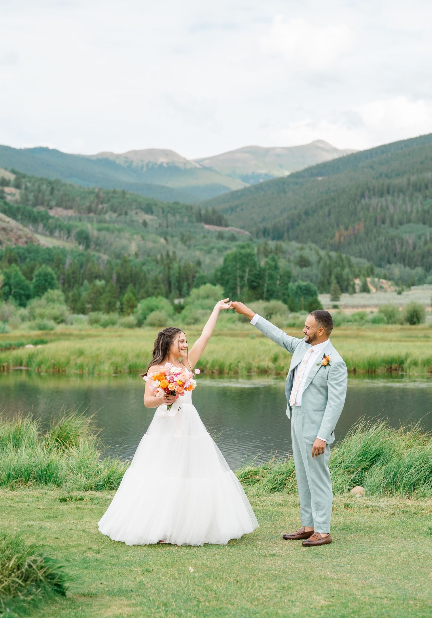 A couple dances together by a mountain lake surrounded by evergreen forests and rolling hills in the background.