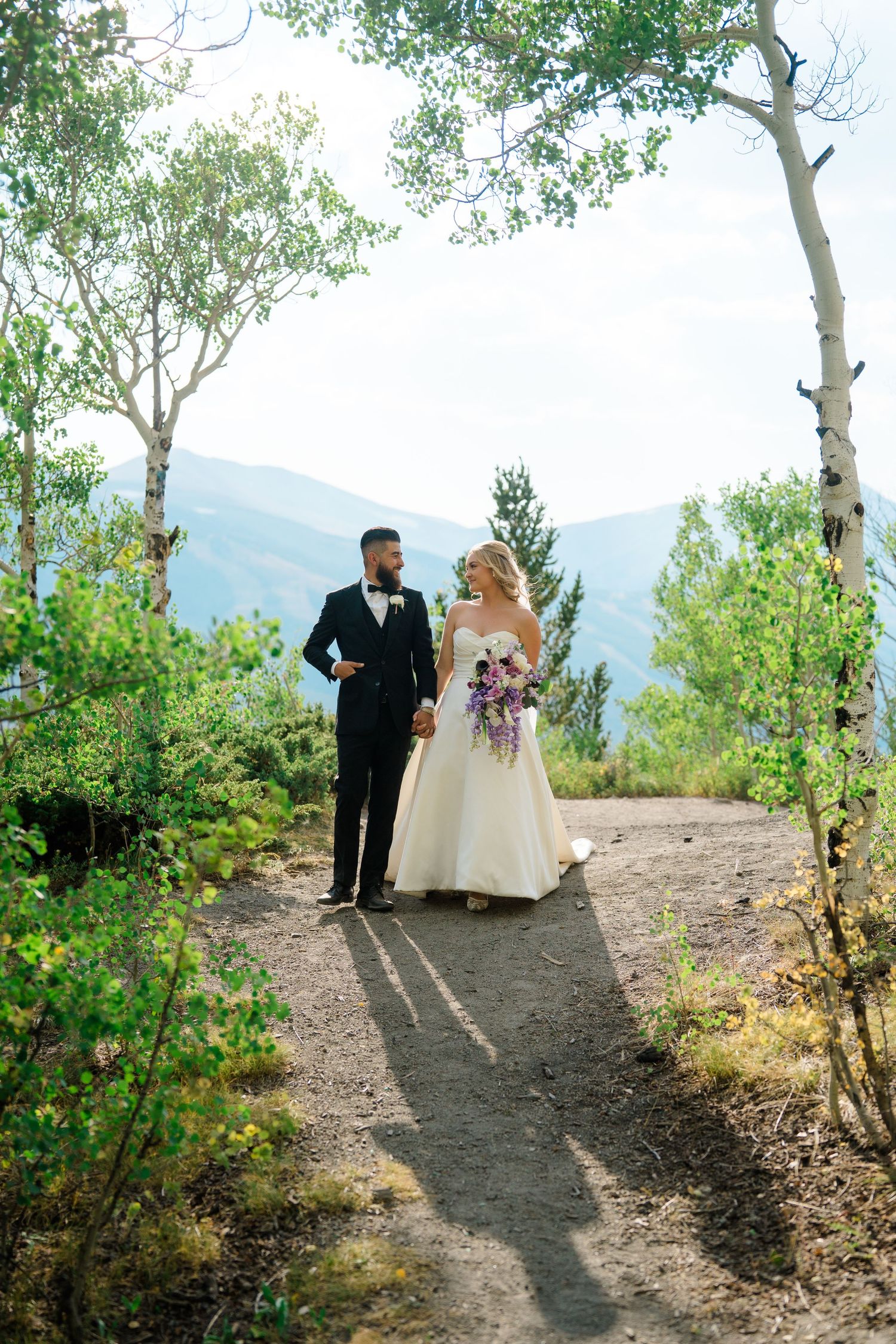 A couple walks along a scenic mountain trail surrounded by aspen trees and natural foliage on their wedding day.
