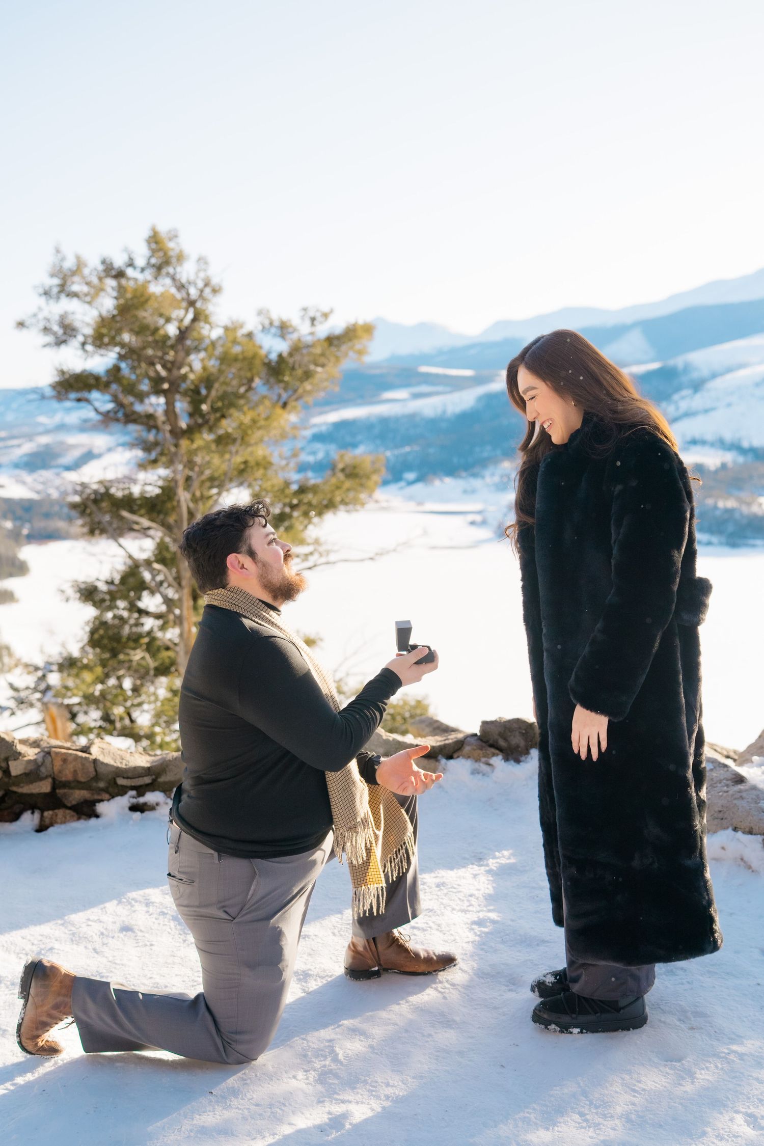 A romantic winter proposal scene with snowy mountains and evergreen trees in the background.