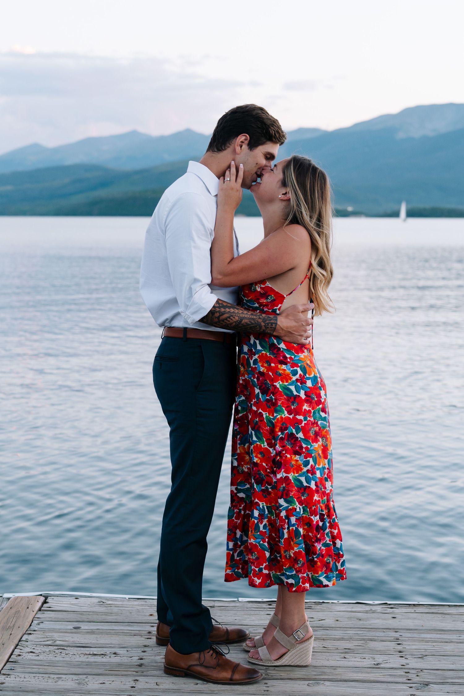 Romantic couple shares tender moments on a wooden dock overlooking a serene mountain lake at sunset.