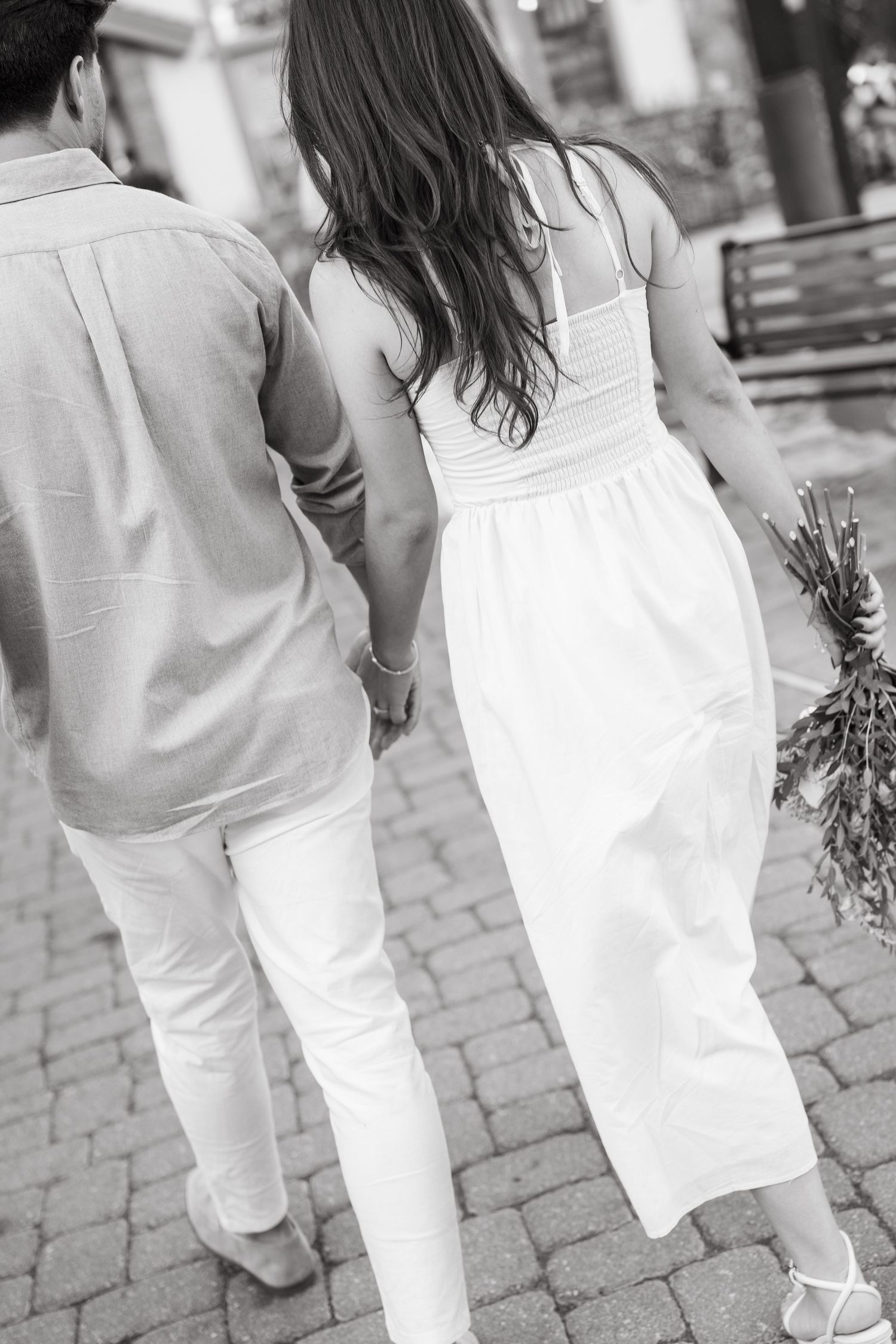 Two people in white clothing walk hand in hand along a cobblestone path in black and white.