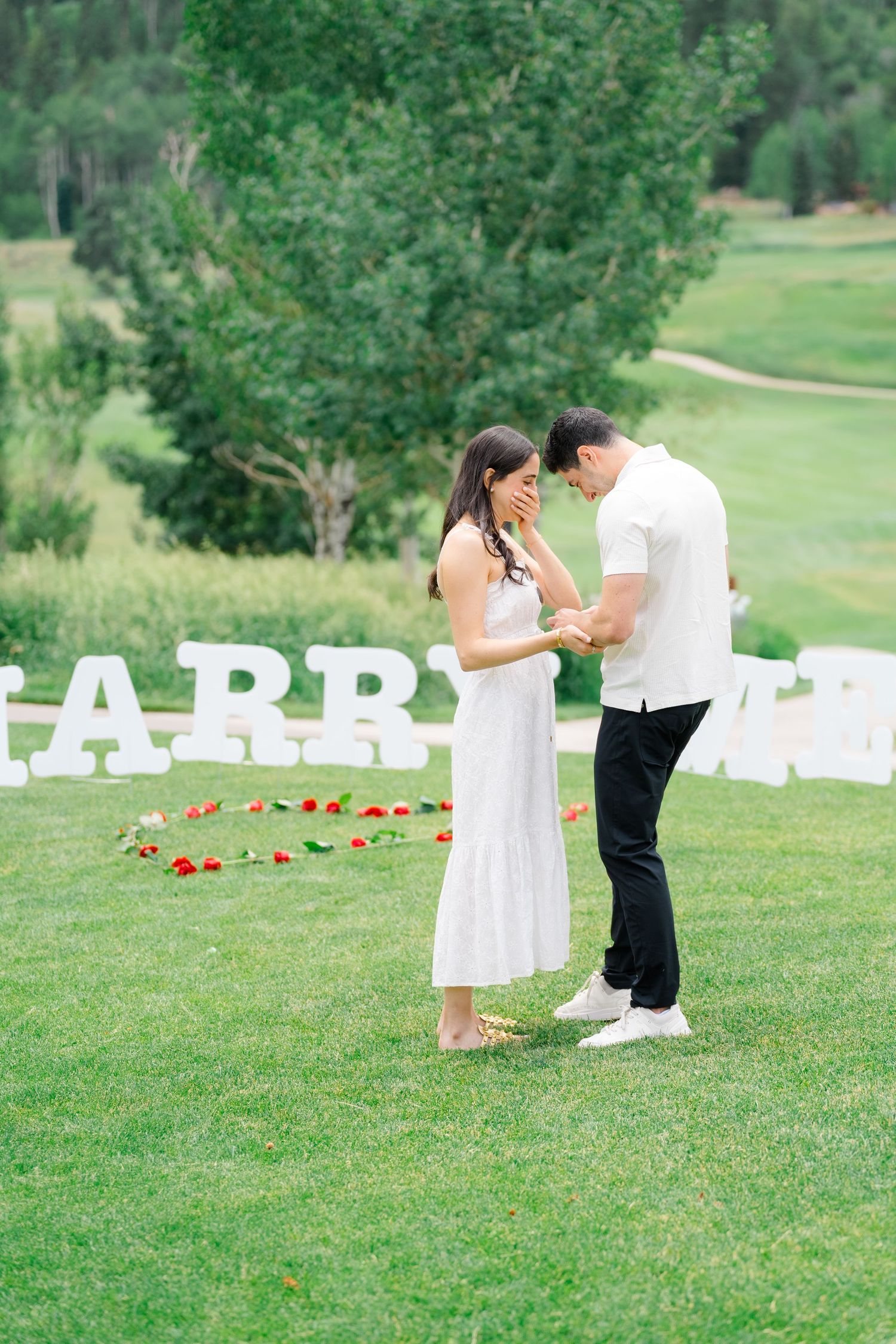 A couple shares an intimate moment during their proposal on a grassy hillside with large white letters in the background.