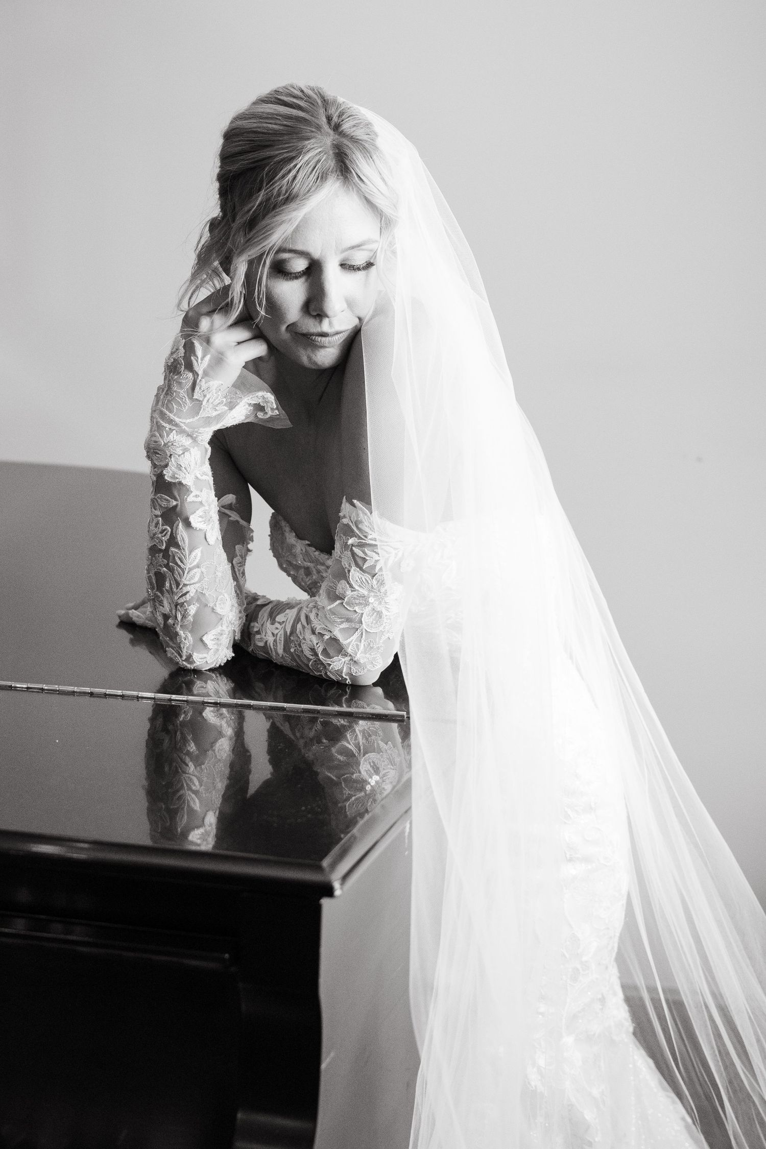 Black and white portrait of a bride in lace sleeves leaning on a piano with flowing veil.