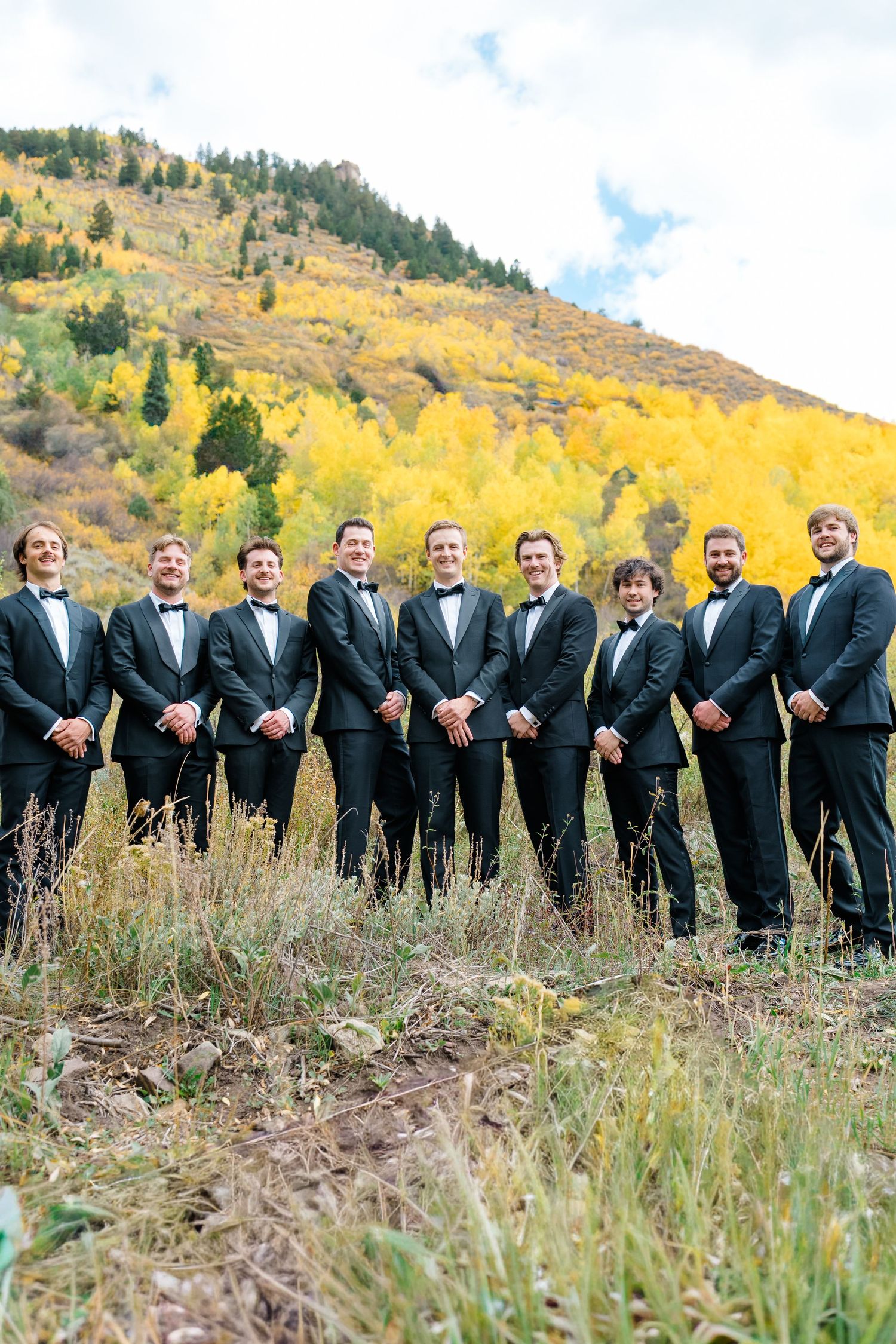 Wedding party members in formal attire stand in formation on a hillside with fall colors in the background.