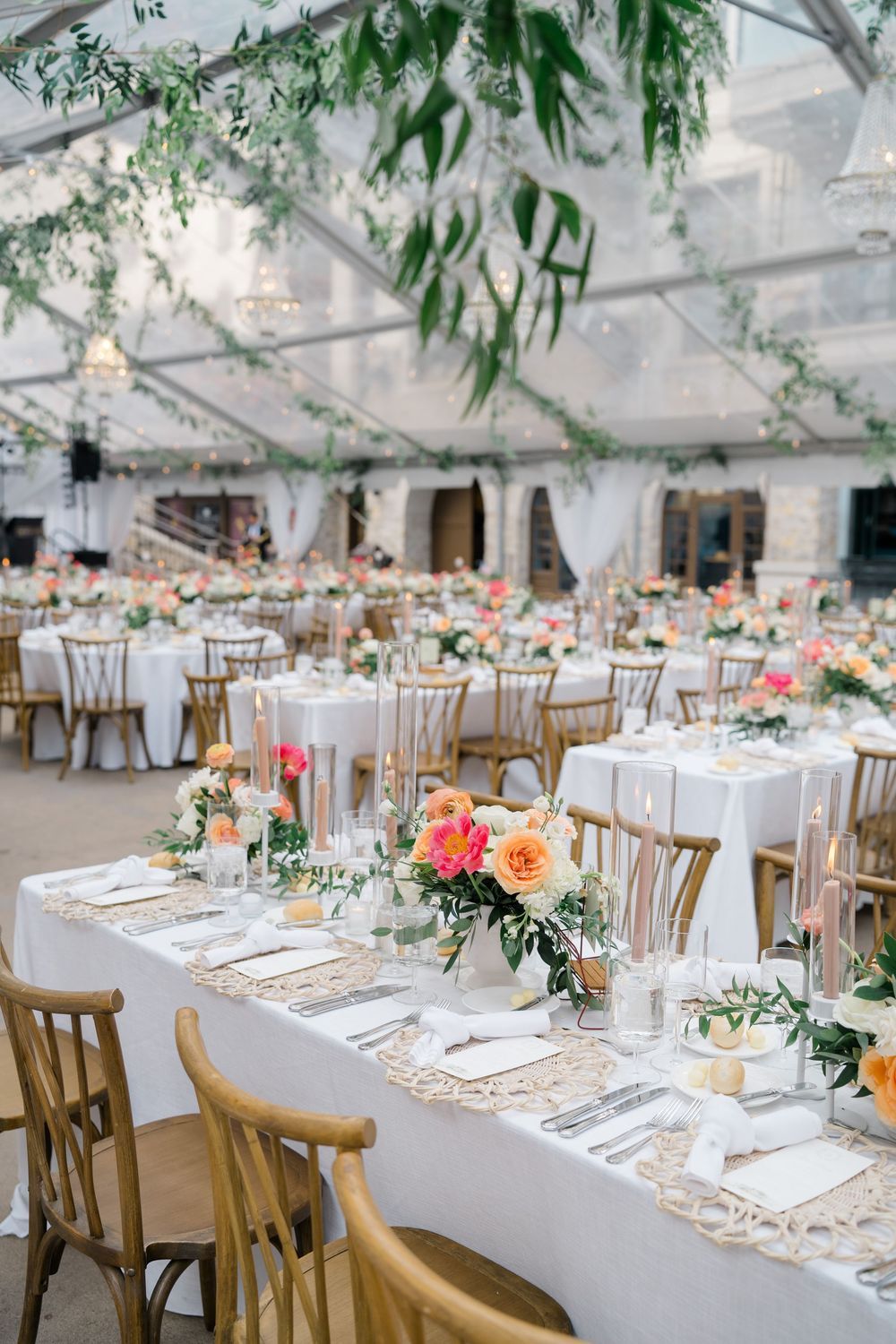 Elegant wedding reception under clear tent with gold chairs, white linens, and vibrant peach floral centerpieces with greenery.