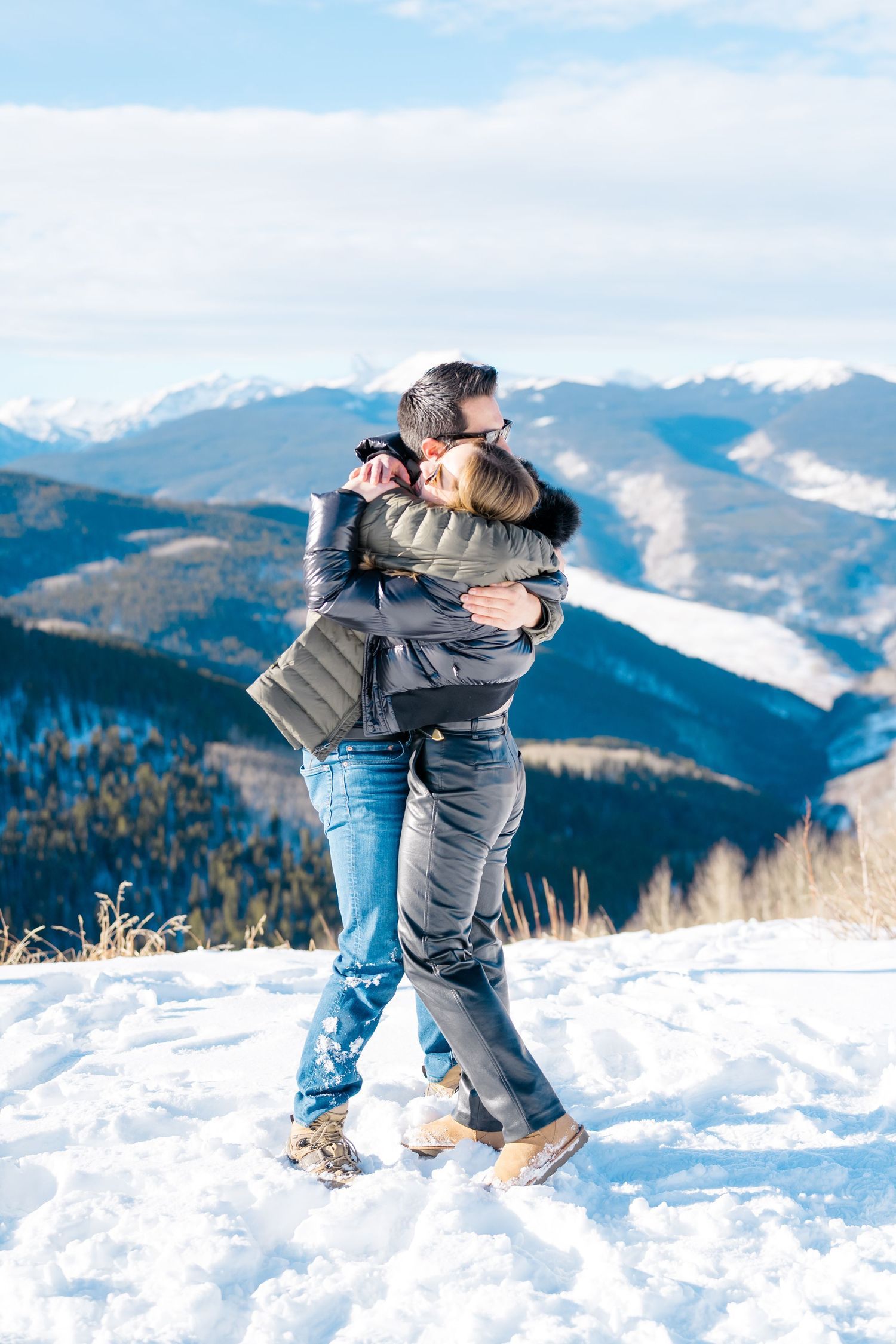 A couple embraces joyfully in the snow with scenic snow-capped mountains and evergreen forests in the background during vail proposal.