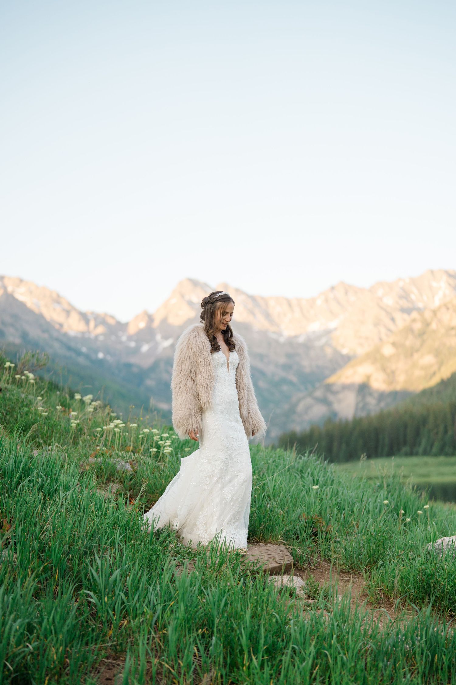 A romantic wedding couple poses against a stunning mountain backdrop with lush green meadows at sunset.