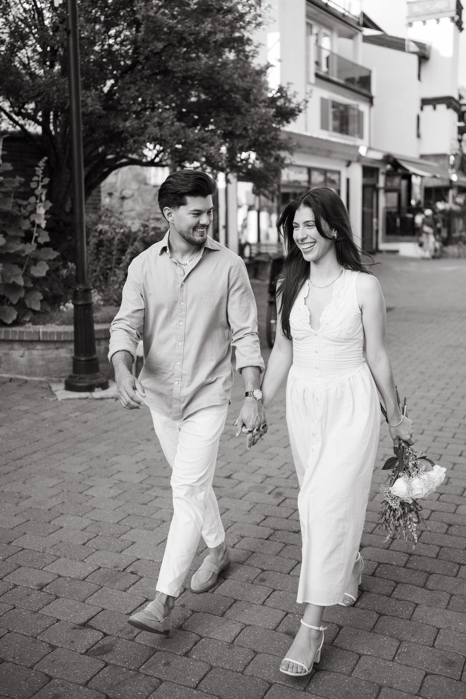A couple in white outfits walks together on a cobblestone street in a black and white photo series.