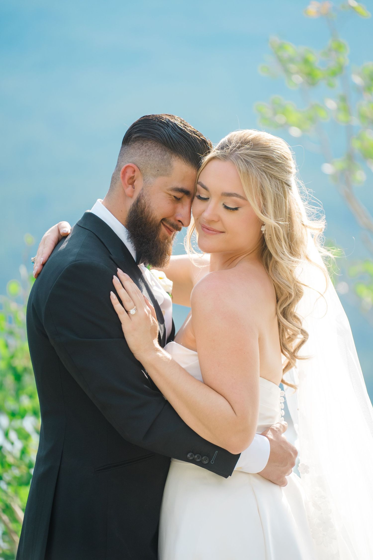 A romantic wedding couple embraces outdoors with sunlit trees and blue sky in the background.