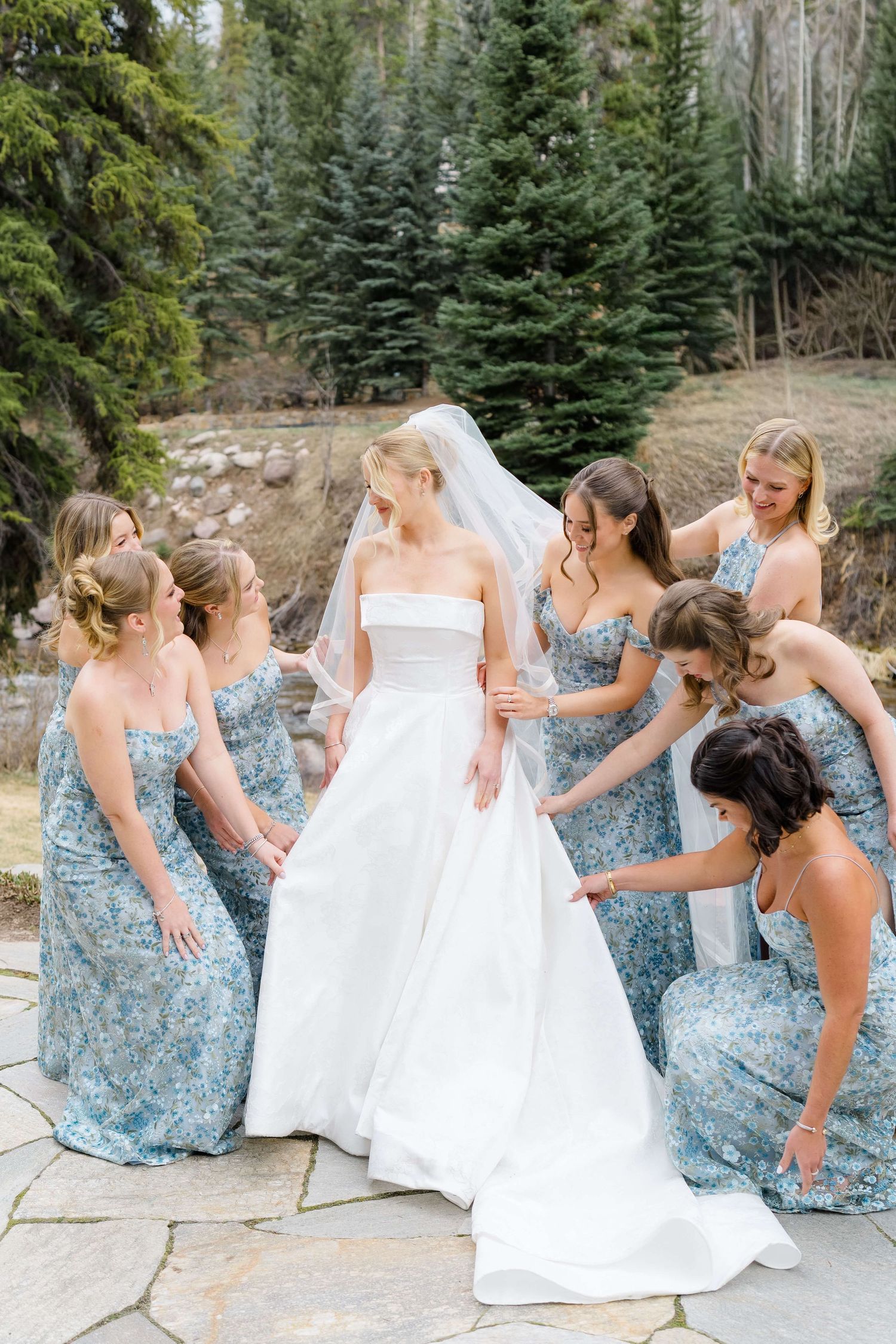Bride in white strapless gown surrounded by bridesmaids in blue floral dresses adjusting her veil outdoors.