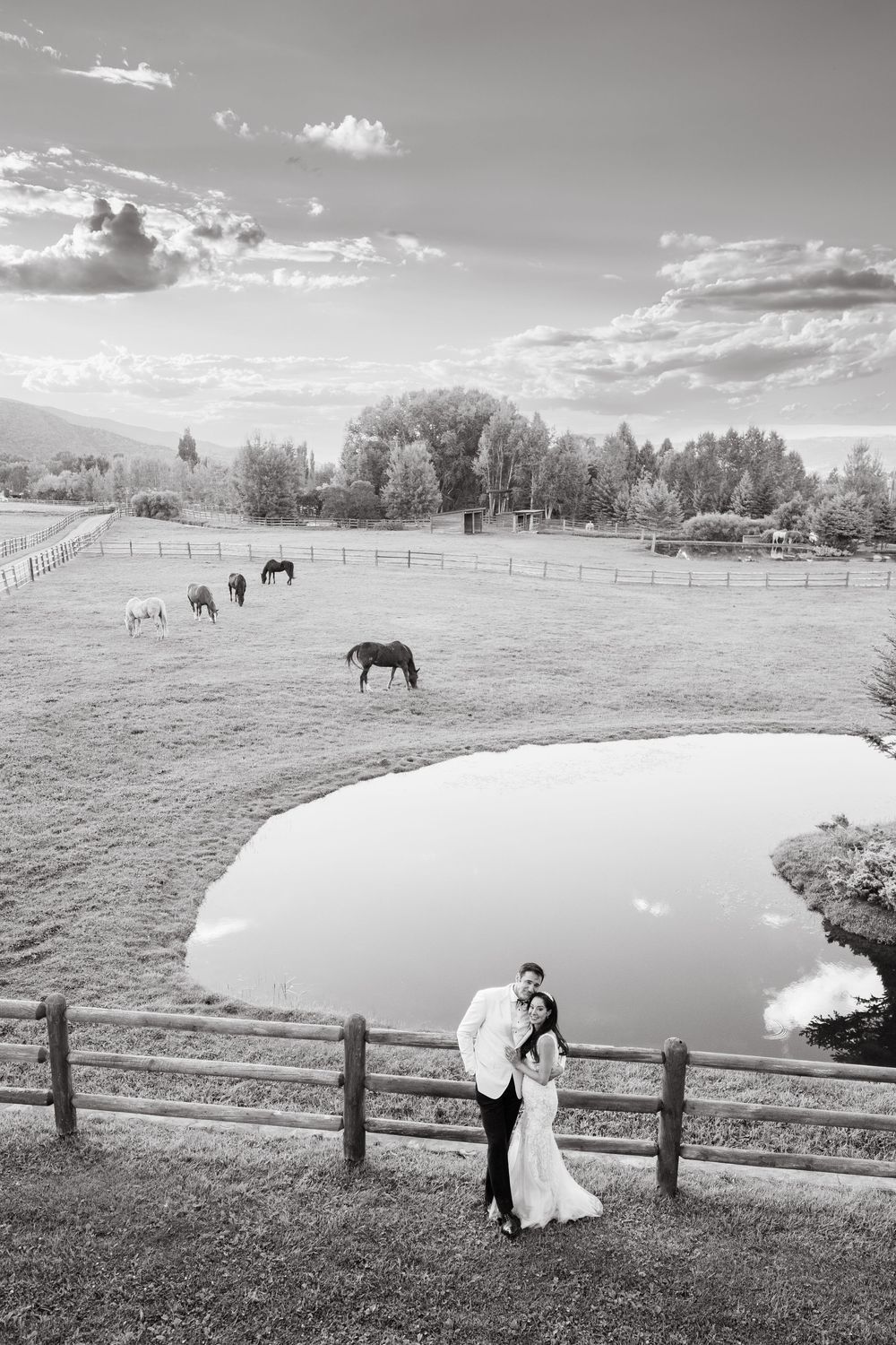 Black and white photo of a bride and groom embracing by a wooden fence overlooking a pond and grazing horses on a ranch.