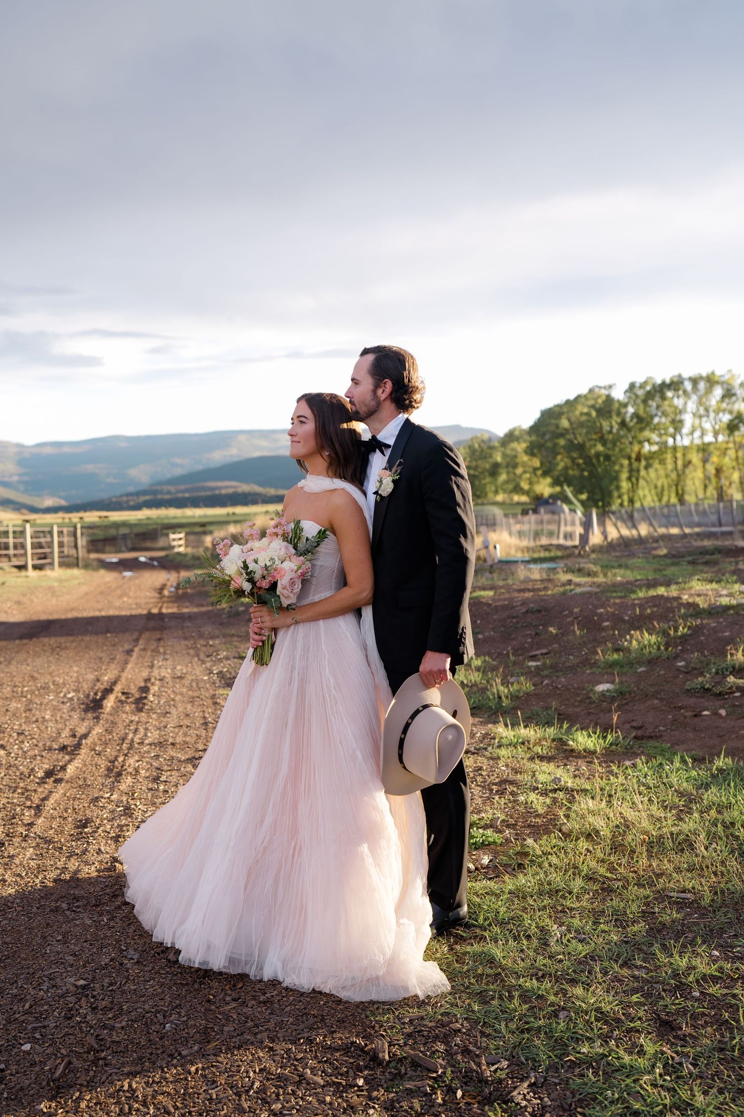 Bride in blush tulle gown holding bouquet stands with groom in black tuxedo holding hat, posing on rural farm with mountain backdrop.