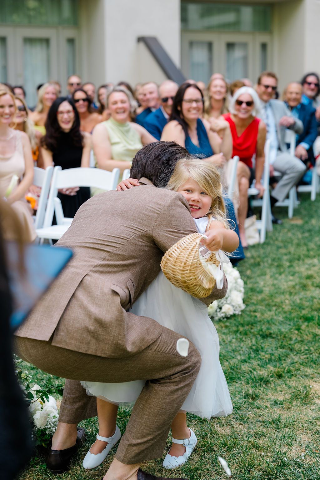 A groom in a tan suit hugs a smiling flower girl during an outdoor wedding ceremony with guests seated behind them.