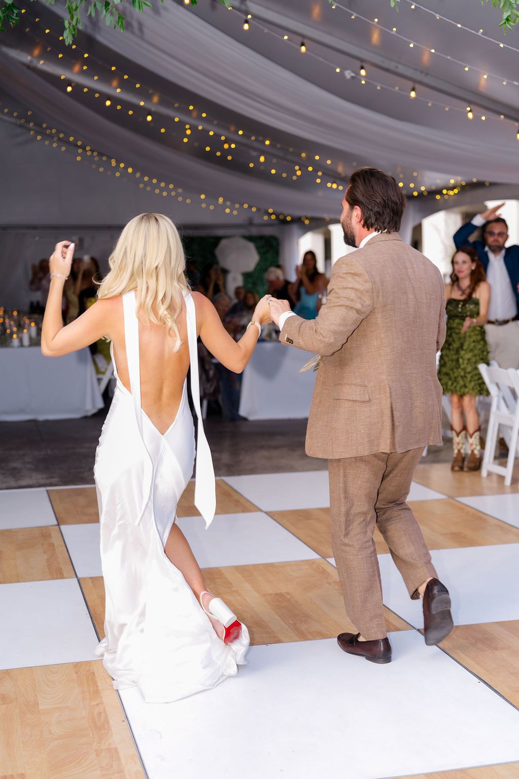 Bride and groom dancing on checkered floor at elegant tented wedding reception with string lights.