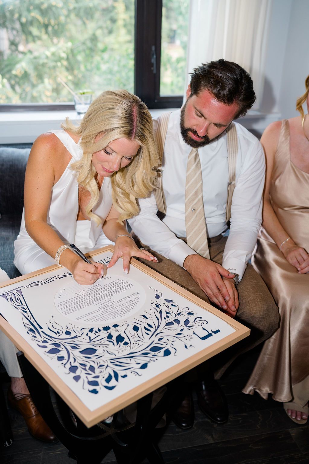 Bride and groom signing their ketubah wedding contract, dressed in white and suspenders at their Jewish ceremony.