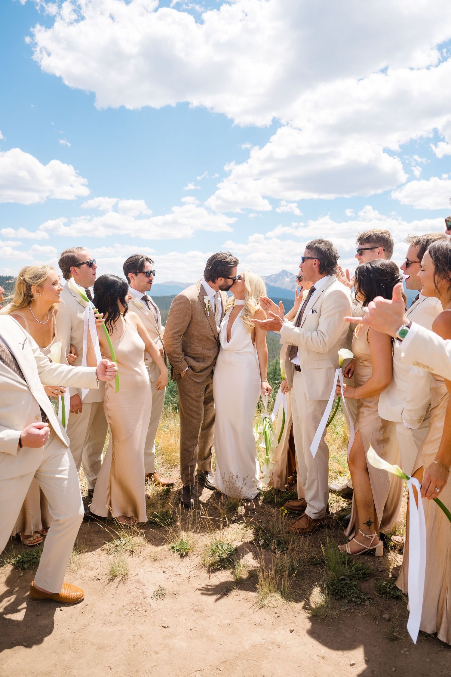 Wedding party in neutral tones celebrating outdoors on a mountain overlook under a bright blue sky.