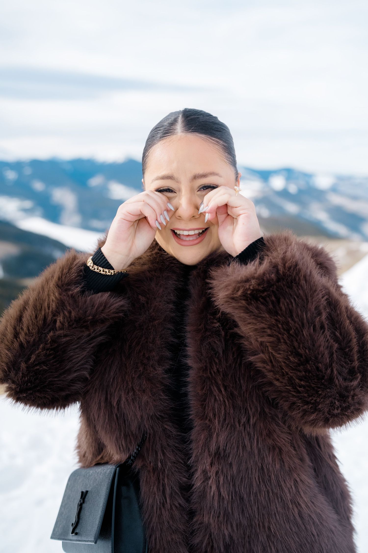 Person in brown fuzzy winter coat smiling and posing playfully against snowy mountain backdrop.