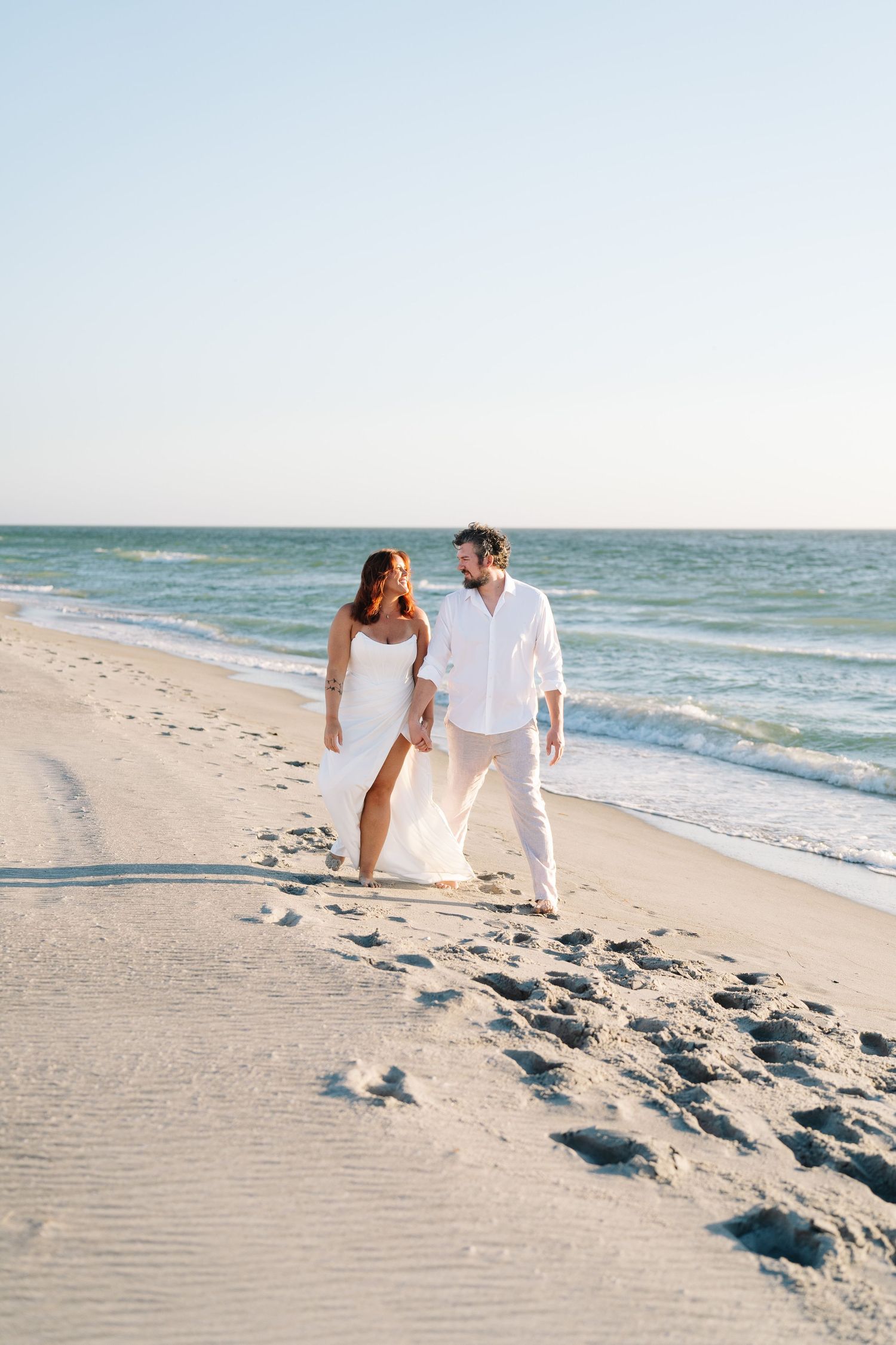 A couple in white outfits sits together on a sunny beach with waves rolling in along the shoreline at sanibel engagement photo session