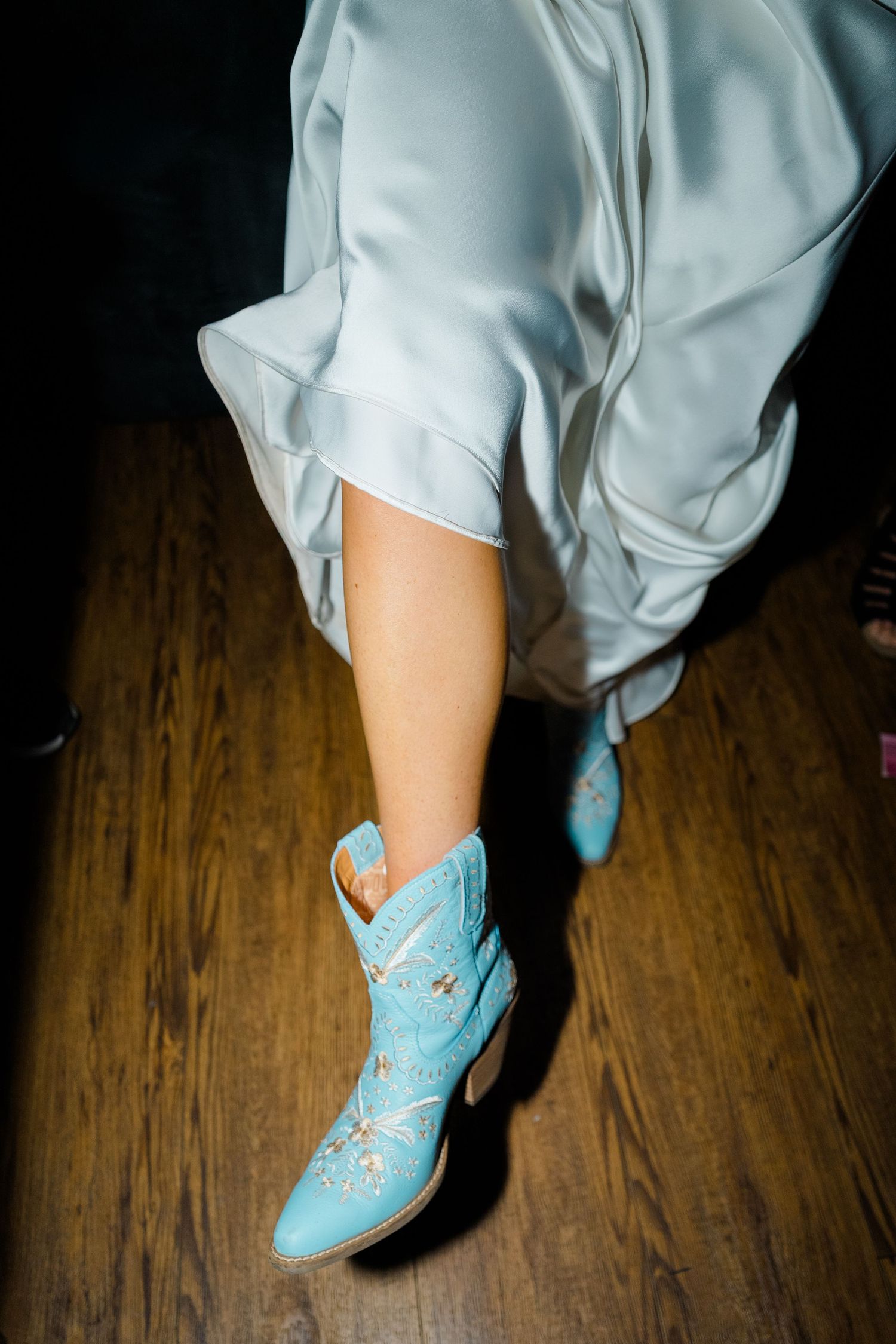 Vail wedding photographer captures Close-up of light blue cowboy boots worn with a flowing white wedding dress on a wooden floor.