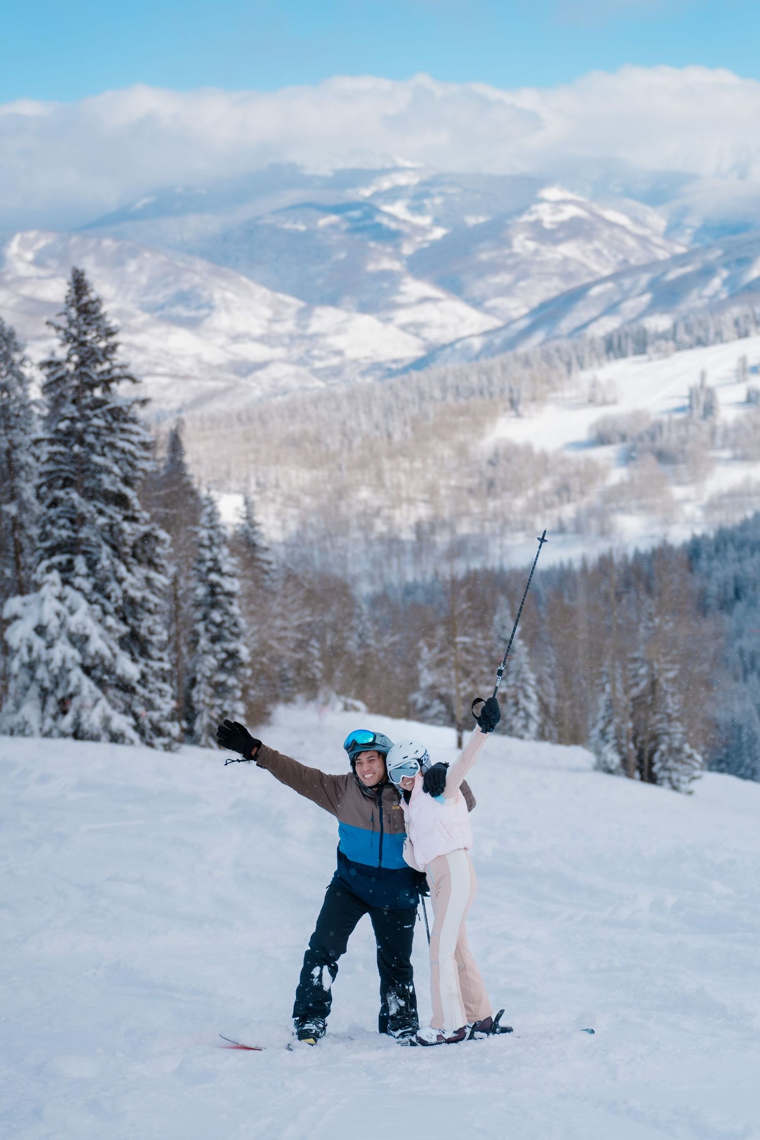 Two skiers celebrate on snowy slopes with scenic mountain views and snow-covered trees in winter landscape.