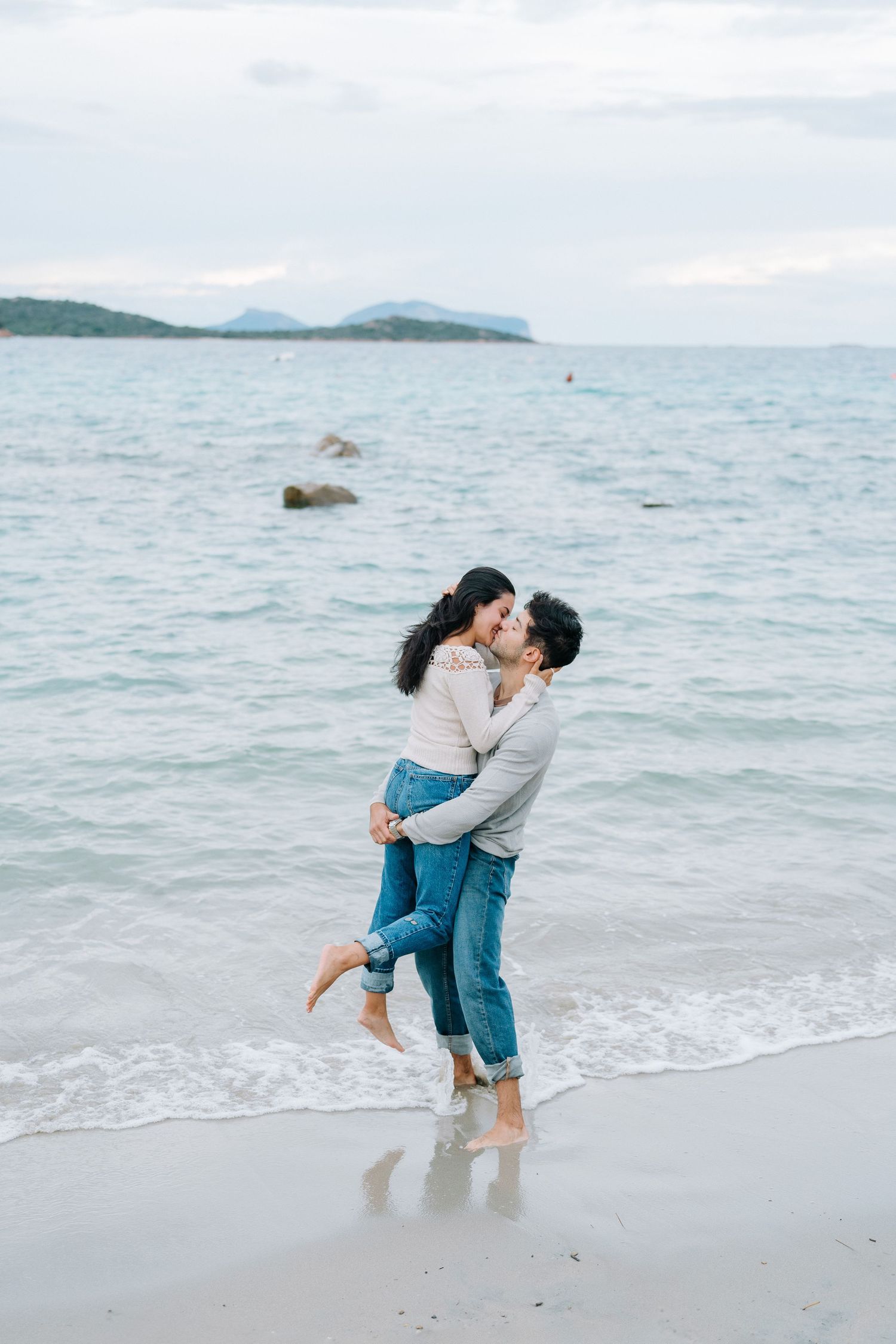 A couple shares romantic moments on a scenic beach with mountains and ocean in the background.