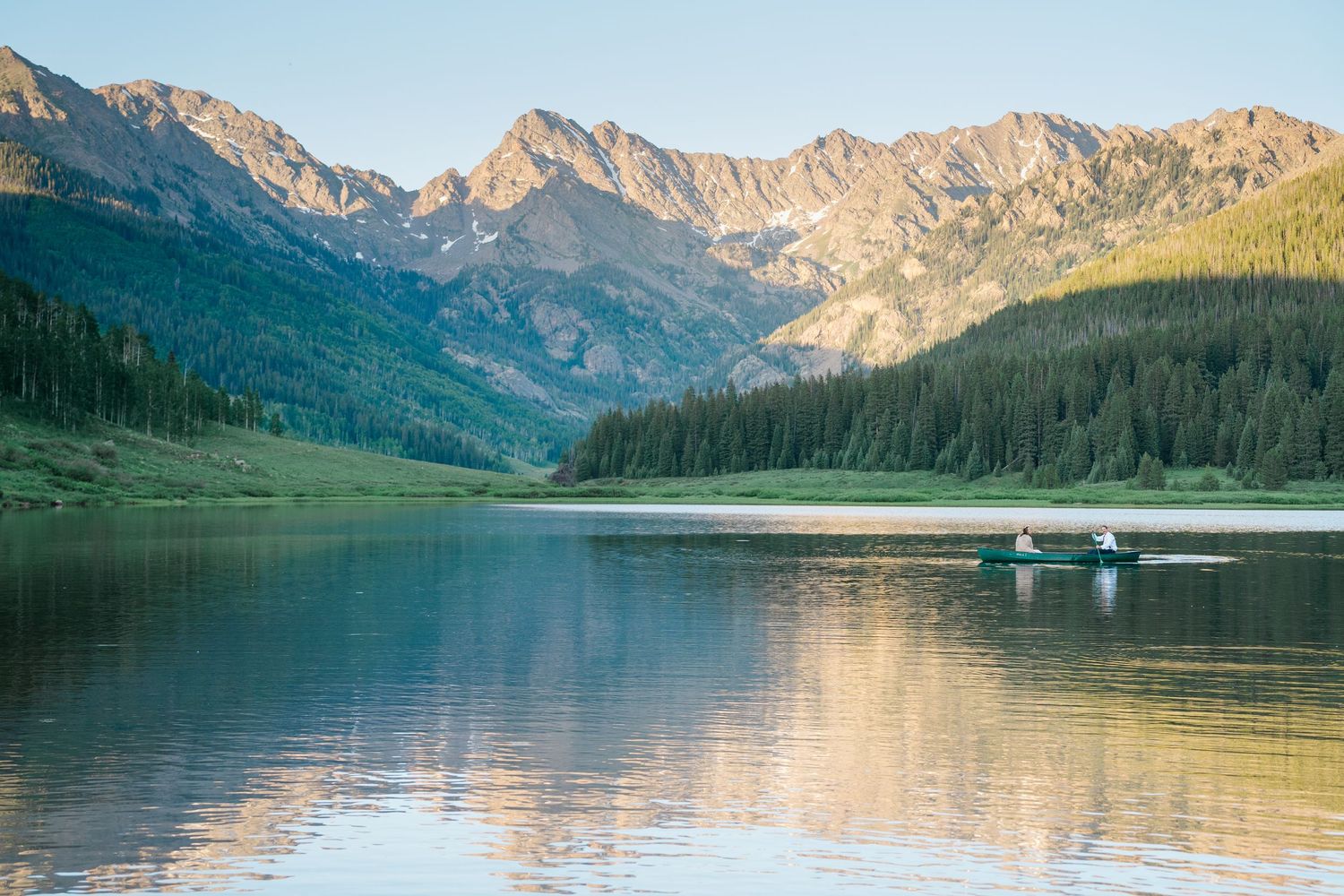 Tranquil alpine lake reflecting rugged mountain peaks and evergreen forests with small boat dock in golden evening light.