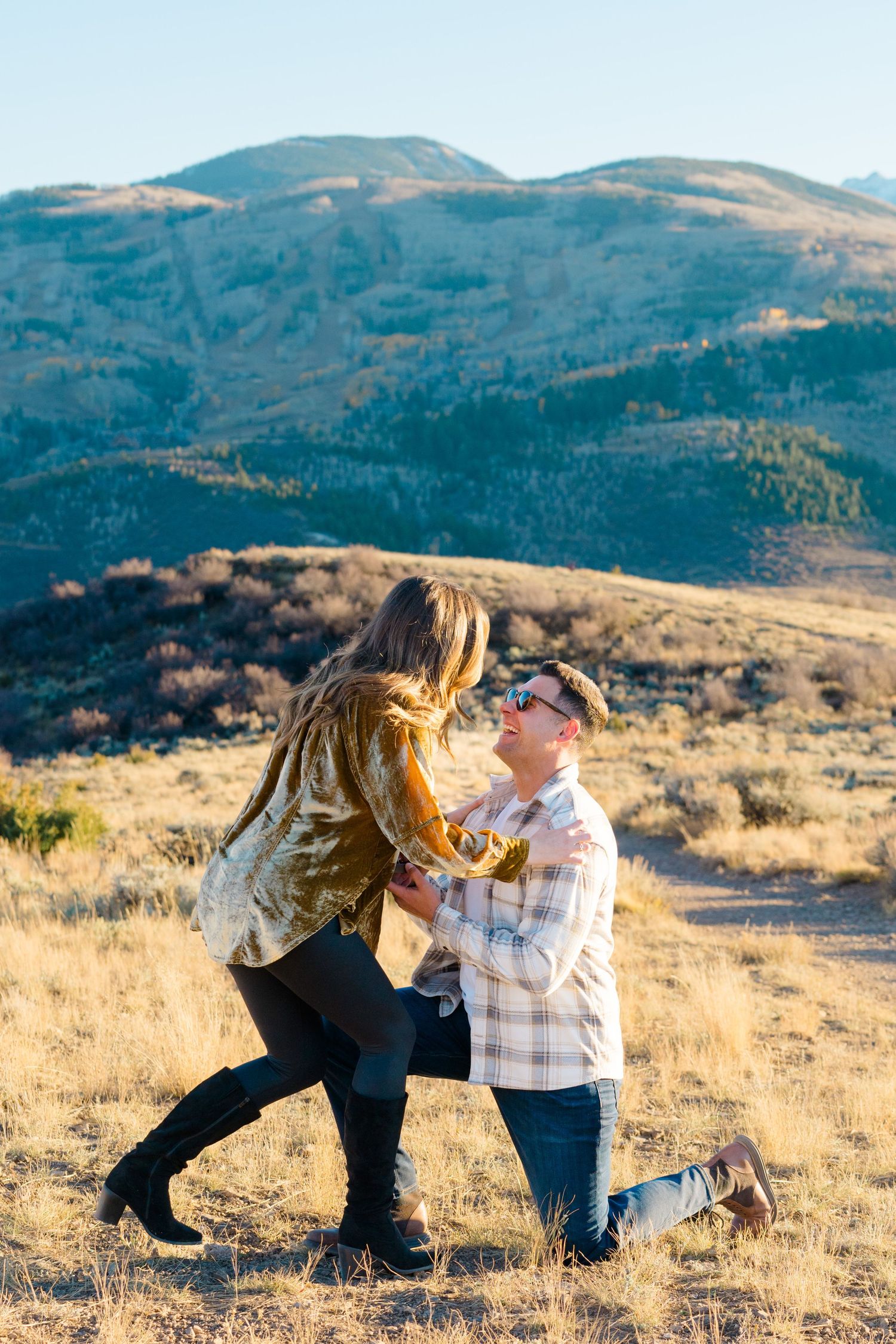 A couple shares a romantic proposal moment on a hilltop with scenic mountain views in the background.