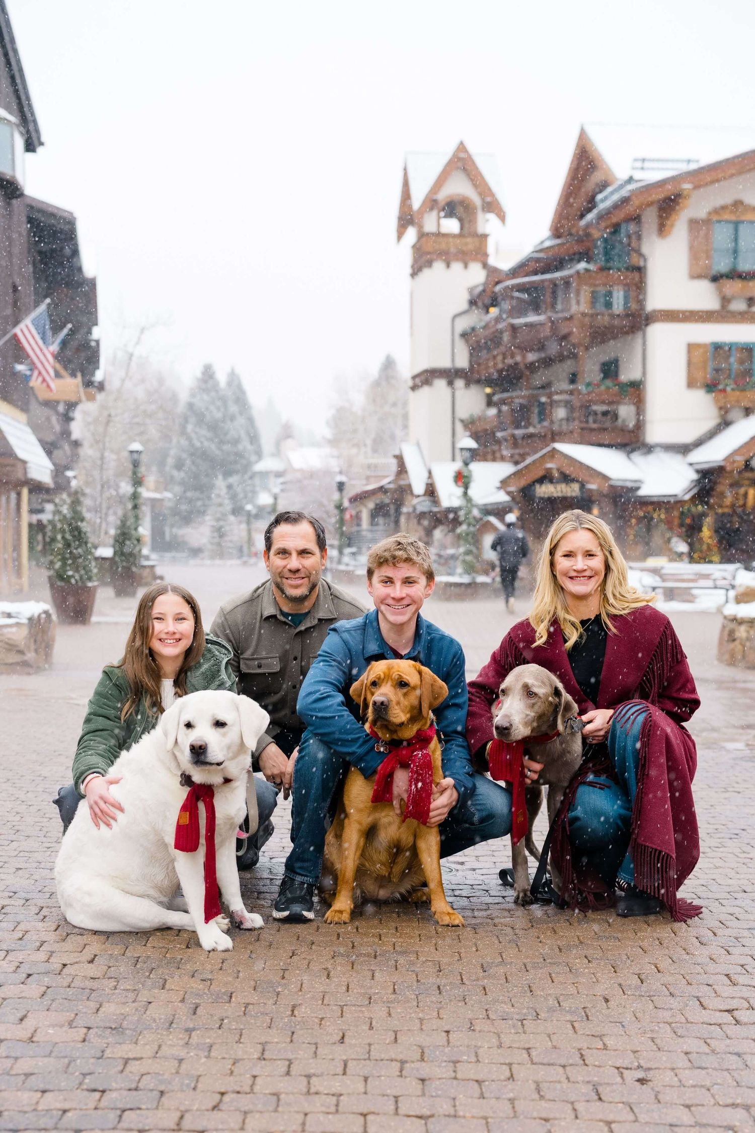 A family and their three dogs pose together on a snowy cobblestone street in front of alpine-style buildings.